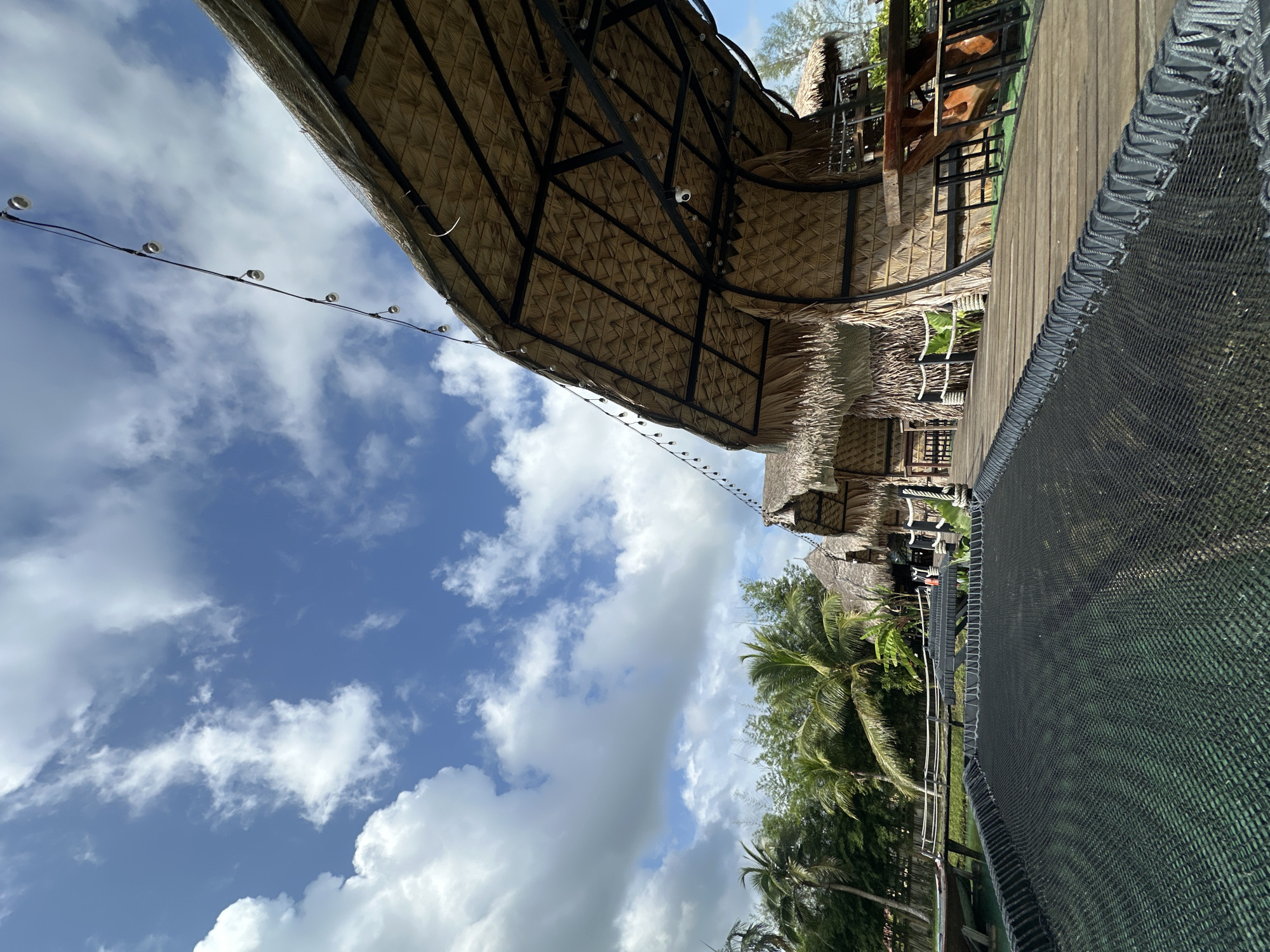 A ground-level, low-angle shot captures a waterfront scene at Tambon Pa Kae Bo Hin, Thailand, under a partially cloudy blue sky. In the foreground, a wide black net, secured by thick grey rope, stretches across the frame. Beyond the net is a wooden boardwalk with a railing. Several thatched-roof structures with woven walls and metal support beams line the boardwalk, some with outdoor seating visible. To the left of the boardwalk, a boat is moored to a wooden pier over calm, greenish water. Lush green vegetation, including palm trees and bamboo, forms a dense backdrop behind the structures. A string of bare light bulbs hangs overhead, illuminated by the daylight.