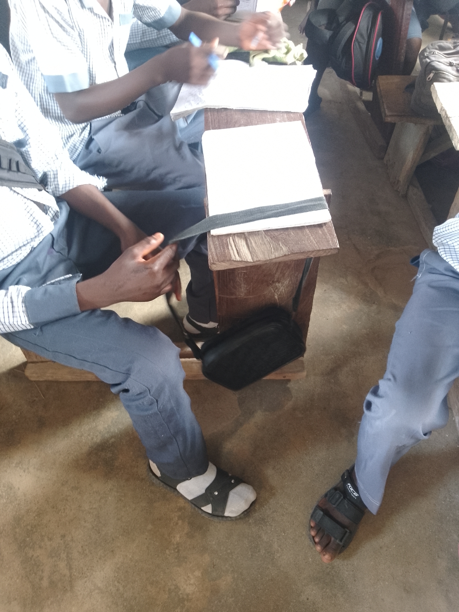 The image shows two individuals, likely students, seated at a rustic wooden desk in what appears to be an outdoor or open-air classroom setting in Serti, Nigeria. One person on the left is wearing a white and blue checkered shirt and grey trousers, with their left hand holding a blue pen and their right hand resting on the desk. The other person on the right is wearing grey trousers and black sandals. Both are focused on their work, with notebooks and a black bag on the desk. The environment is simple, with a dirt floor and basic wooden furniture, suggesting a rural or under-resourced educational facility. The lighting indicates it is daytime.