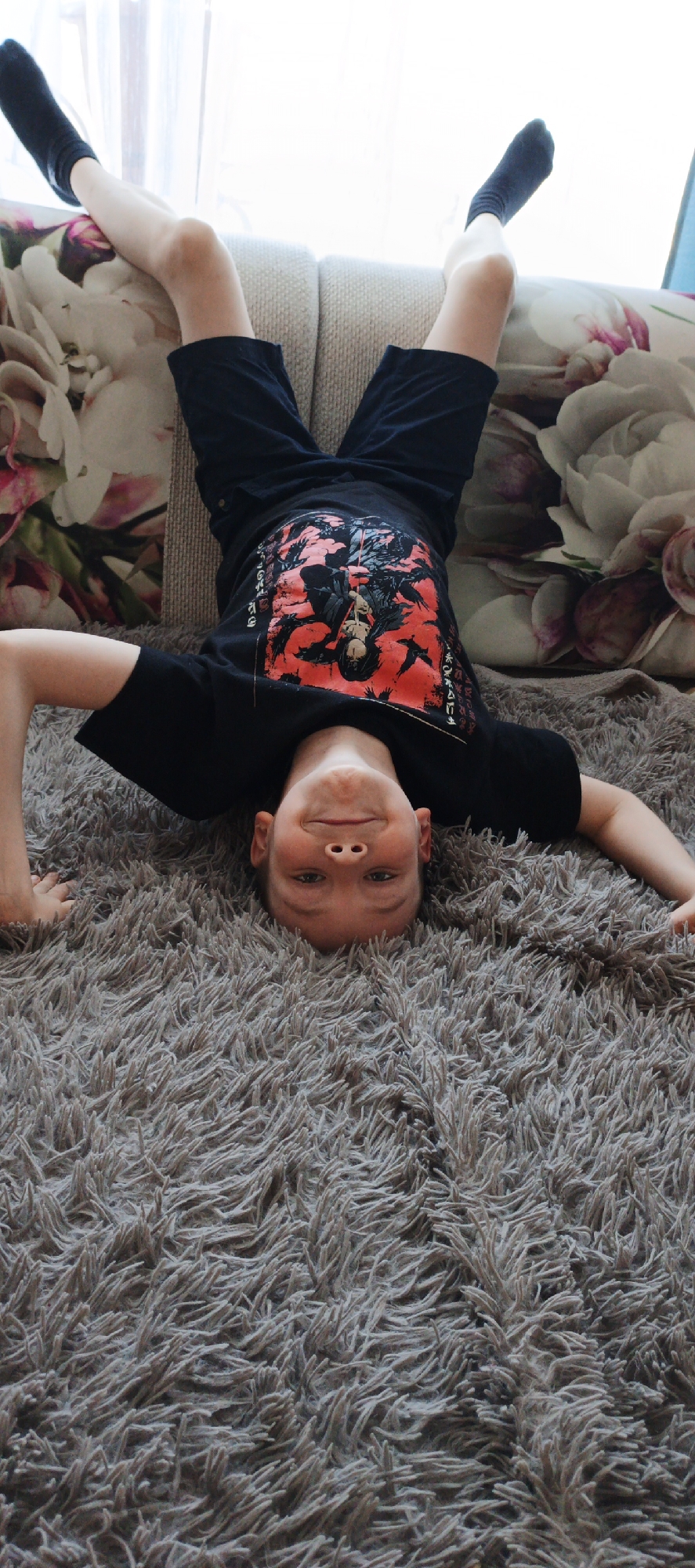 A boy is lying upside down on a gray shag rug. His legs are propped up on a couch, which is covered with pillows featuring a floral pattern. He is wearing black socks, shorts, and a t-shirt with a red and black graphic. His arms are outstretched on the rug, and he is looking towards the camera with a slight smile. Based on prior context, the photo was taken in Chaikovsky, Russia.