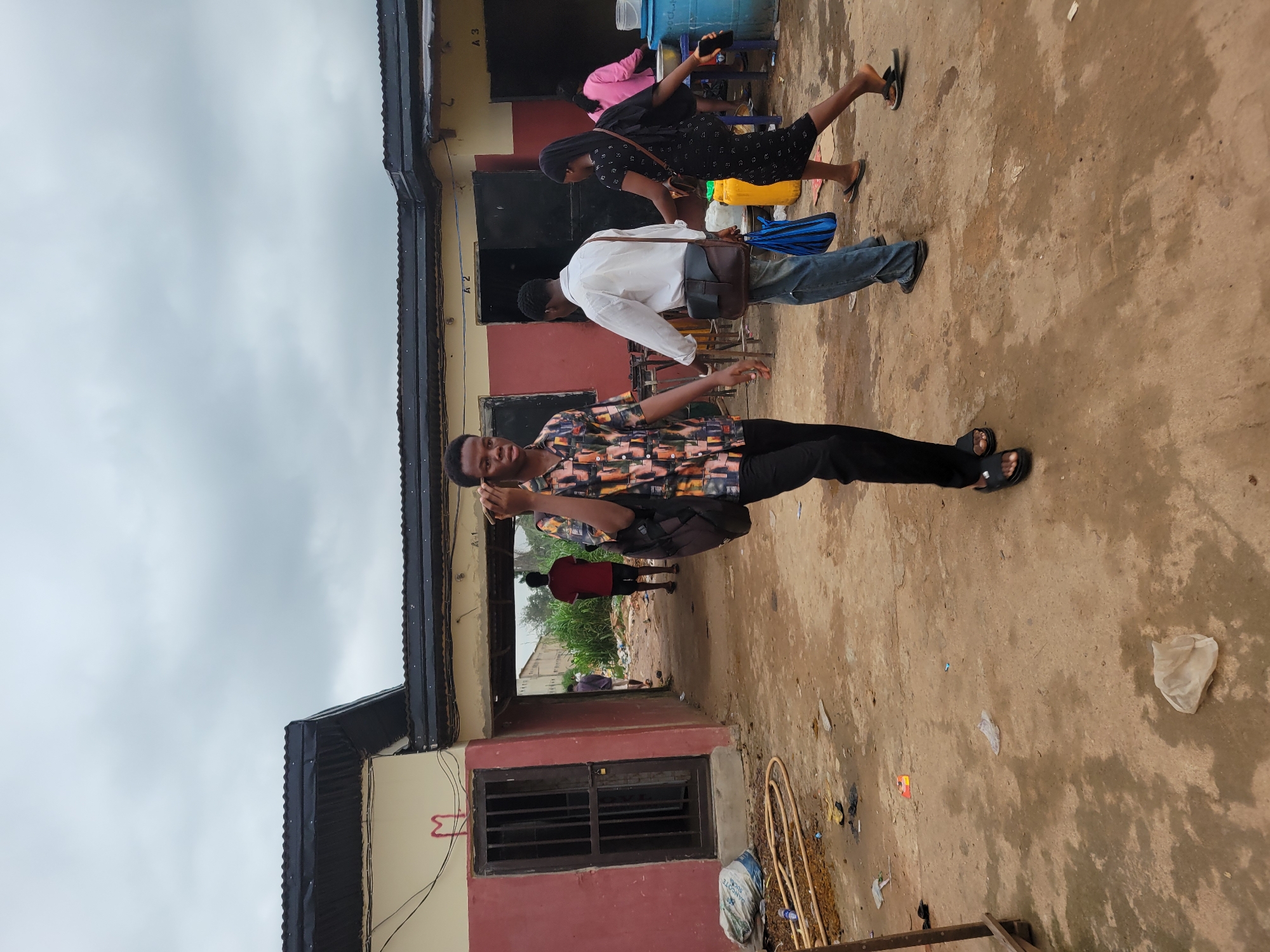 In Egbagu, Nigeria, a young Black male is captured in a medium shot, walking towards the camera while holding a mobile phone to his ear. He is wearing a short-sleeved, multicolored patterned shirt and black pants. He carries a black backpack slung over his right shoulder. The background reveals a somewhat disheveled courtyard with a red and beige building featuring dark metal window coverings and doors. Several other people are visible in the background: a Black male in a red shirt and shorts walks away from the camera; another Black male in a white shirt and blue jeans, carrying a brown satchel, walks past; and a Black female in a black dress with a white pattern, also carrying a bag, walks away from the camera. A Black female in a pink top and dark pants is seen near some large blue and yellow containers. The ground is made of packed dirt with scattered debris, including plastic bags and what appears to be a yellow hose. The sky is overcast with gray clouds. The overall scene suggests a busy outdoor area, possibly a market or a communal space, with individuals engaged in their daily activities.