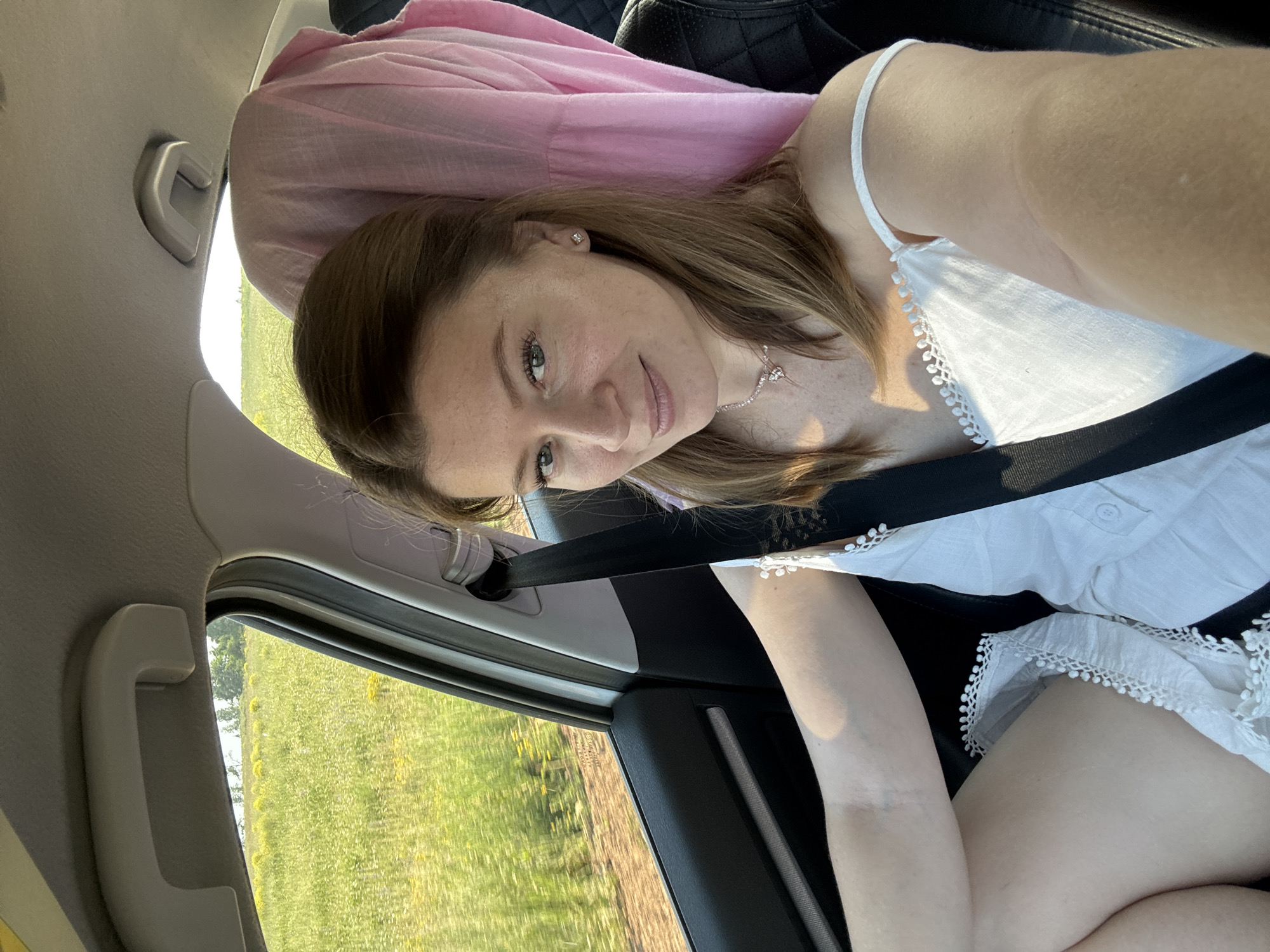 A woman is taking a selfie in the passenger seat of a car, which is moving through a rural area. She is looking at the camera with a slight smile and has her seatbelt on. The car's interior is visible, including the dashboard, door, and a pink fabric draped over the headrest. Outside the window, a field of green and yellow wildflowers is visible, suggesting the setting is likely the countryside. The image does not provide enough information to determine if the location is in the city of Van'ki, Russia, or any other specific city.