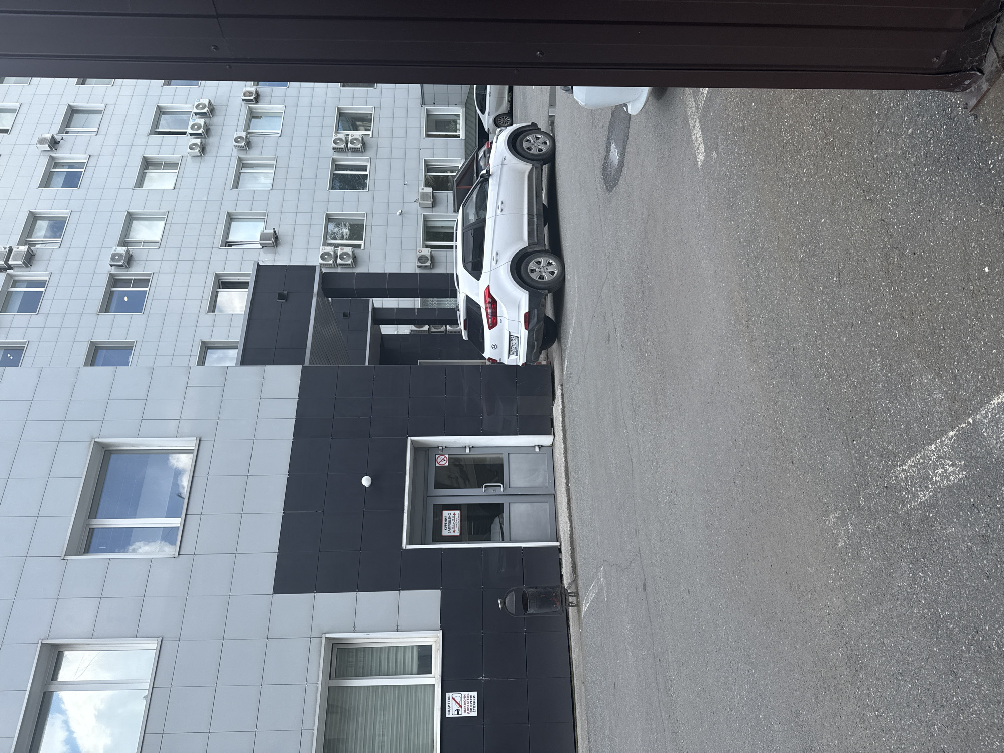 The image depicts the exterior of a modern, multi-story building with a grey, tiled facade. The building has numerous windows and air conditioning units mounted on its exterior. In the foreground, there is an asphalt-paved area that appears to be a parking lot or courtyard.
A white Hyundai Creta SUV is parked in the foreground, facing to the right. The vehicle has a license plate that reads "H232TC 159". Behind the white SUV, another car, a silver sedan, is partially visible. To the left of the white SUV, there is a dark-colored trash receptacle. A set of double glass doors is visible on the lower level of the building, with a "no smoking" sign and other signage in Russian posted on the doors.
Two additional signs are visible on the wall to the left of the entrance: one with a car icon and text in Russian, indicating "DRIVER! TURN OFF THE ENGINE AT THE PARKING LOT!", and another smaller sign with a crossed-out cigarette icon and text. The general environment suggests an urban setting, likely a commercial or administrative complex. The license plate suggests the location is in Perm, Russia, as indicated by the "159" region code.