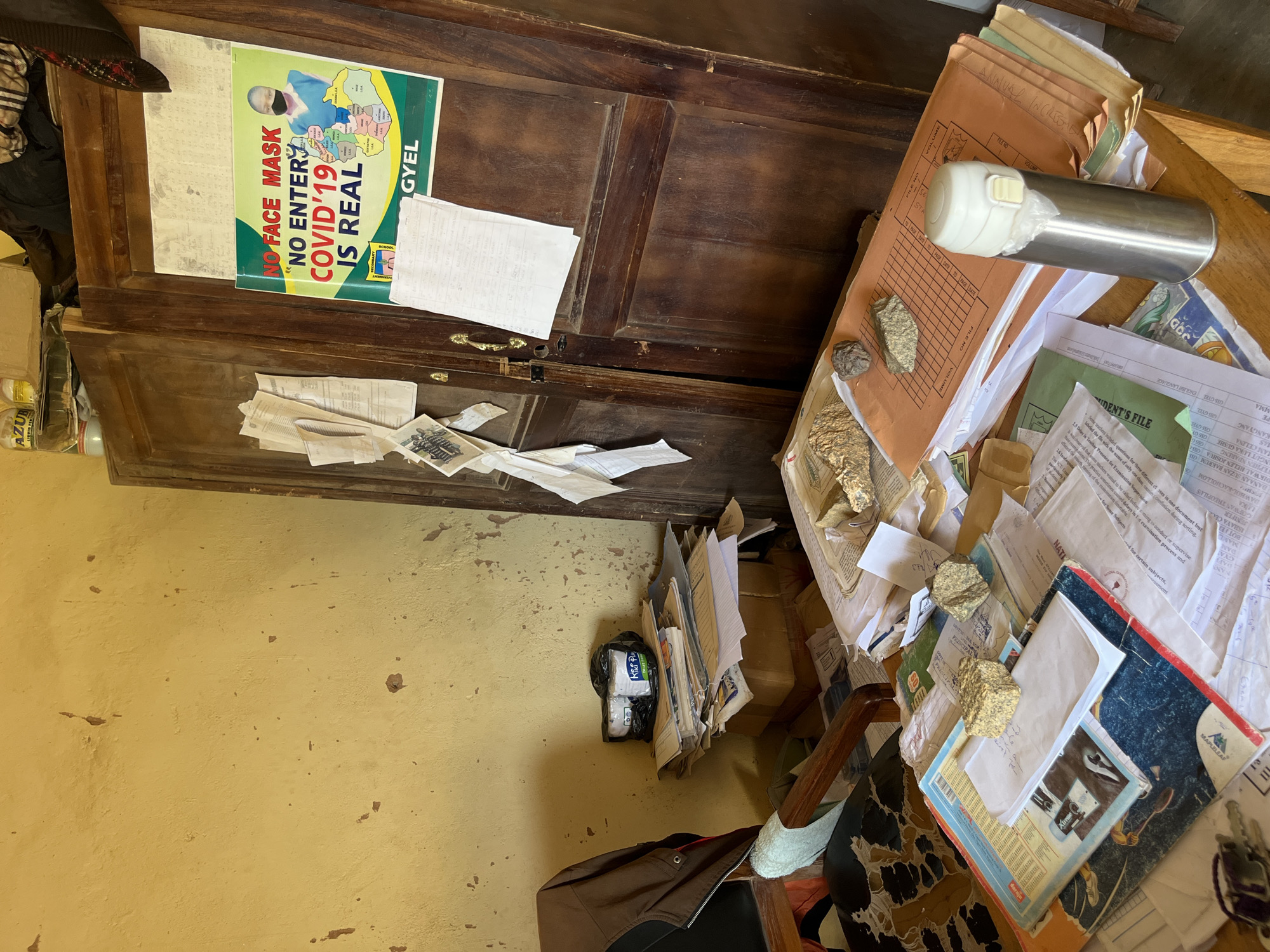 The image depicts an interior scene, likely a room or office, with a cluttered wooden desk in the foreground. The desk is covered with stacks of papers, folders, and various objects. A metallic thermos is prominently displayed on a stack of orange folders. Two rocks or geological samples are placed on top of some documents. To the left, a chair with a patterned seat is partially visible.
Behind the desk, a large, dark wooden cabinet or wardrobe stands against a yellow, textured wall. A poster with the text "NO FACE MASK NO ENTER COVID'19 IS REAL" and an image of a person wearing a mask is affixed to the cabinet door. Several loose papers are also attached to the cabinet.
On a small table to the left of the cabinet, there are more papers and two white cylindrical objects wrapped in black plastic. The overall impression is one of disarray and the presence of informational materials related to public health, specifically COVID-19. The location is identified as Bukuru, Nigeria.