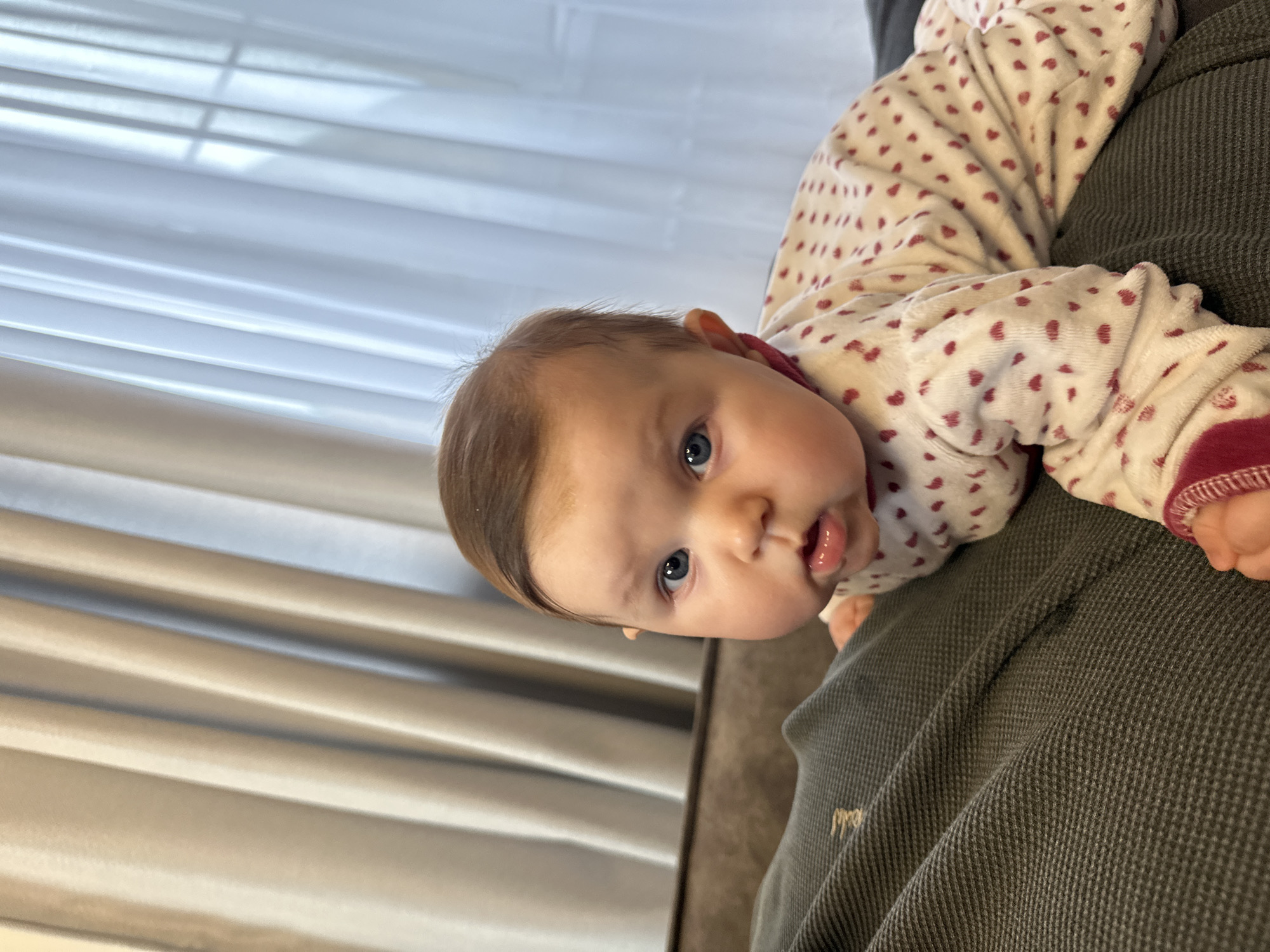 A baby with blue eyes and light brown hair lies on a person's lap, wearing a white and red heart-patterned pajama. The person's garment is dark grey. In the background, blinds cover a window. The location of the image is not possible to determine.
