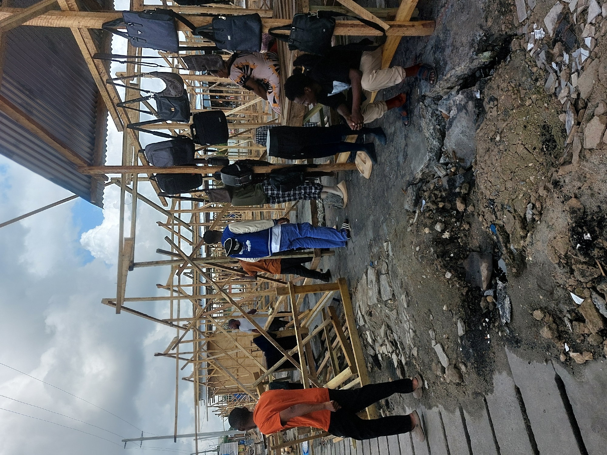 The image captures an outdoor market scene in what appears to be Dar es-Salaam, Tanzania, under a cloudy daytime sky. Several people, mostly men, are present amidst makeshift wooden stalls used for displaying merchandise.
The primary subjects are the people and numerous dark-colored bags, including shoulder bags, briefcases, and possibly backpacks, which are hung from the wooden structures. Some individuals are seen interacting with the display, while others are standing, sitting, or walking through the area. One person in an orange shirt is prominent in the foreground, walking away from the camera. Another person in a black top and light trousers is seated, looking down at their hands.
The setting is characterized by temporary-looking wooden scaffolding forming shelves and a partial roof covered with corrugated metal sheets. The ground is uneven and appears to be a mix of packed dirt, loose soil, gravel, and rubble, including broken concrete pieces. This suggests an unpaved, informal market environment. Power lines are faintly visible against the sky in the distance.
Notable details include the casual attire of the people, consisting of everyday clothing like t-shirts, jeans, and sandals. The natural lighting indicates it is daytime, with a bright sky despite significant cloud cover. The overall scene depicts a bustling, functional market area. No discernible text is clearly visible on any items, clothing, or signage.