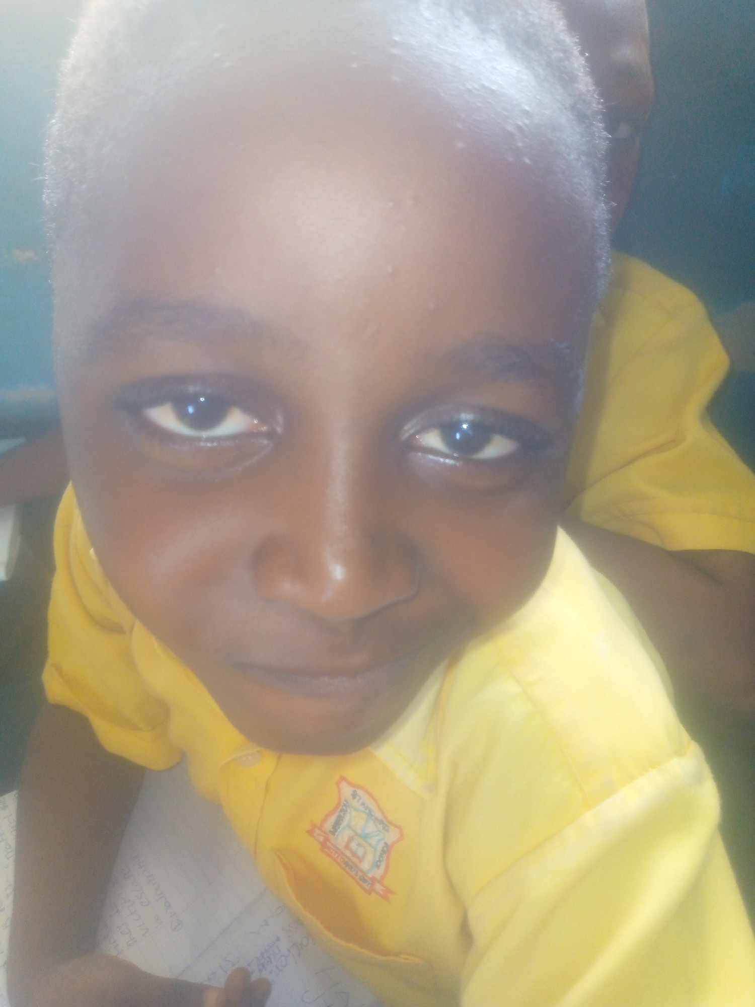 A close-up, slightly angled photograph shows a young Black child wearing a yellow collared shirt with a logo on the left side. The child is indoors, with the edge of a blue wall visible at the top left. Part of another person in a similar yellow shirt is visible behind the child. In the foreground, there is a piece of paper with handwritten text. The lighting is bright, possibly from artificial sources. The location is inferred to be in the city of Ikorodu, Nigeria, based on the prompt.