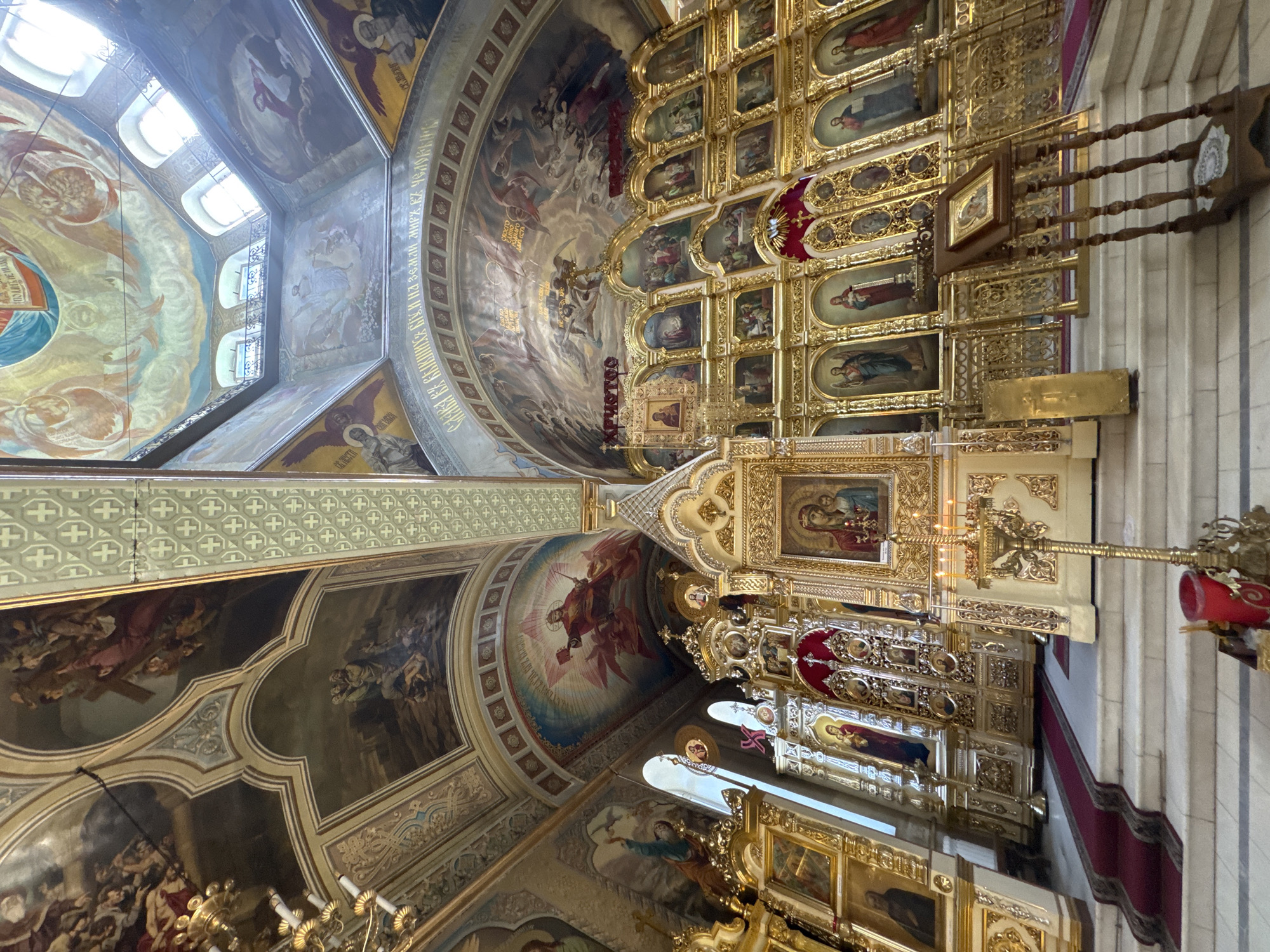 The image depicts the interior of an ornate church, likely an Eastern Orthodox church, showcasing a grand iconostasis and elaborately decorated ceilings. The iconostasis, a wall of icons, is gilded and adorned with numerous religious paintings and carved elements. Several icons are visible, including depictions of the Virgin Mary and Child, Christ, and various saints, rendered in traditional Byzantine style.
The ceiling is a canvas of religious murals, featuring biblical scenes and figures, painted in vibrant colors with golden accents. The architectural style suggests a historical church, with vaulted ceilings and arched windows. Natural light streams in from windows, illuminating the interior and highlighting the golden ornamentation.
In the foreground, a red carpet leads up a set of white marble steps towards the iconostasis. A brass candelabra with lit candles stands to the right of the steps, adding to the sacred atmosphere. A wooden stand with an icon is placed on the steps, indicating ongoing religious observance. The overall impression is one of solemnity, grandeur, and deep spiritual significance. The specific location is not identifiable from the image itself.