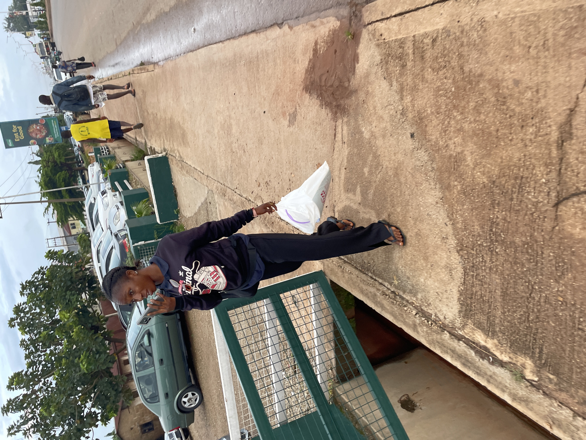 A woman walks on a sidewalk in Jos, Nigeria, holding a white plastic bag in her right hand and a phone to her ear with her left hand. She is wearing a black sweatshirt with a graphic design and black pants. She has black braided hair and is wearing dark flip-flops. Behind her, there are parked cars and trees, and further in the background, there is a billboard that reads "Eat for Good." The sidewalk is made of concrete and appears wet in some areas. Other people are visible walking on the sidewalk and road in the distance.