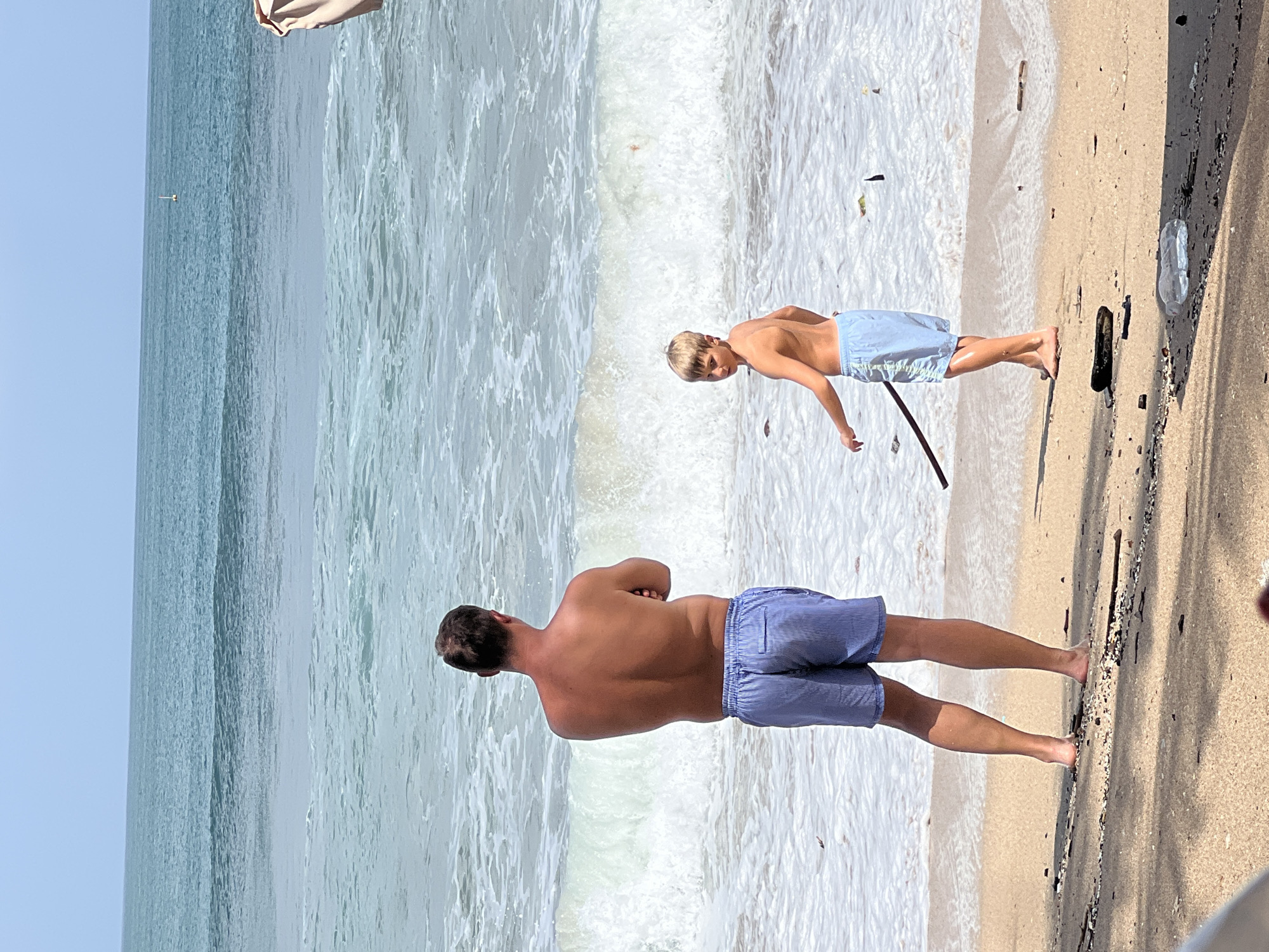 Two males are standing on the beach at Kuta, Indonesia, with the ocean in the background. The male on the left, an adult, is facing away from the camera, looking out at the ocean with his arms crossed. He is wearing blue striped swim trunks. The male on the right, a child, is facing away from the ocean and towards the adult. He is looking back at the camera and holding a stick. He is wearing blue swim trunks. The ocean is a light blue-green with white waves. The sand is a light brown color. The sky is clear and blue.
