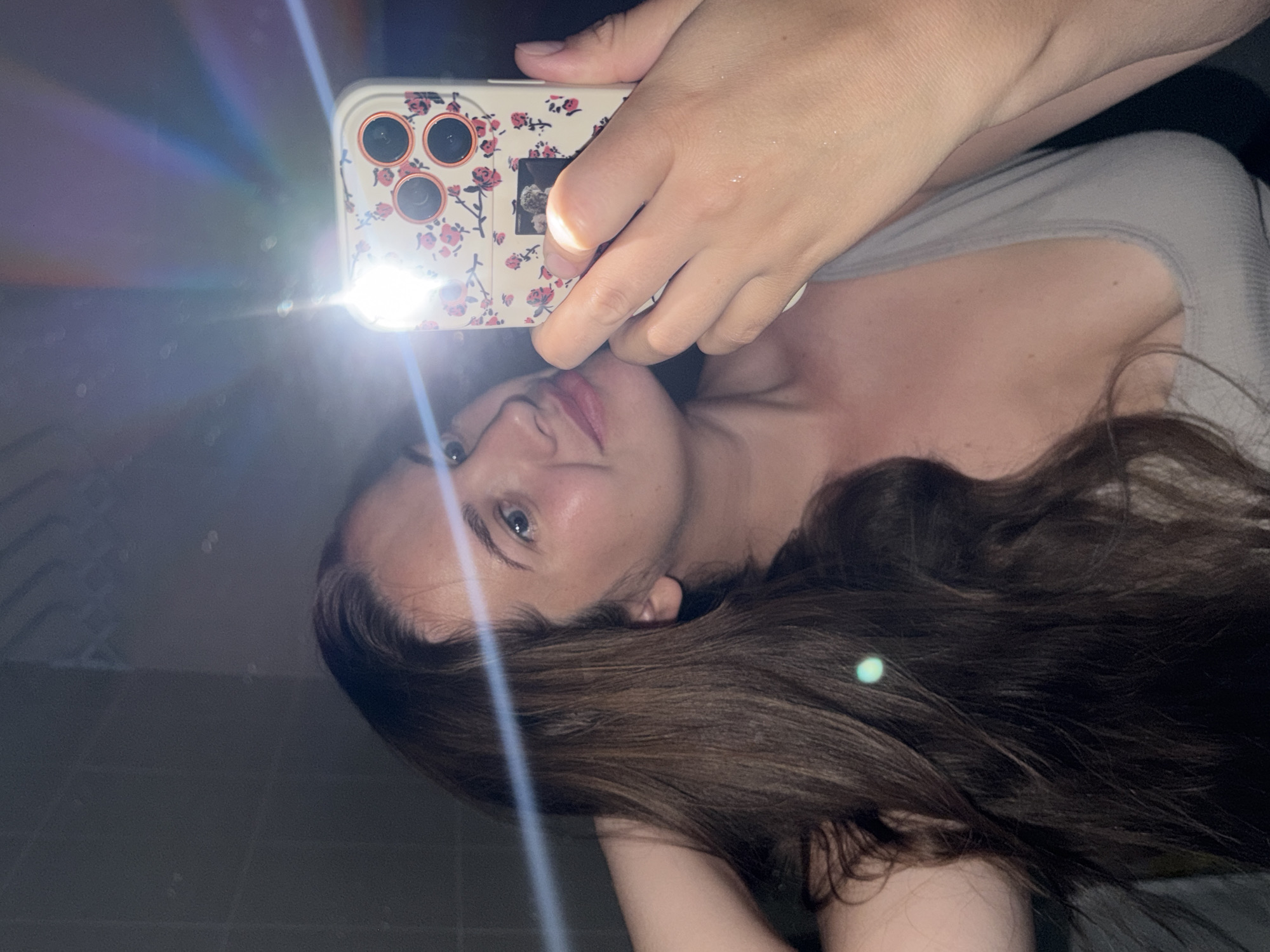 A woman is taking a selfie in what appears to be a dimly lit bathroom with tiled walls. She has long, brown hair and is wearing a light-colored tank top. She is holding a smartphone with a floral case in her right hand, and the flash is illuminating her face and the surrounding area, creating lens flares. The woman is looking at the phone's reflection in a mirror.