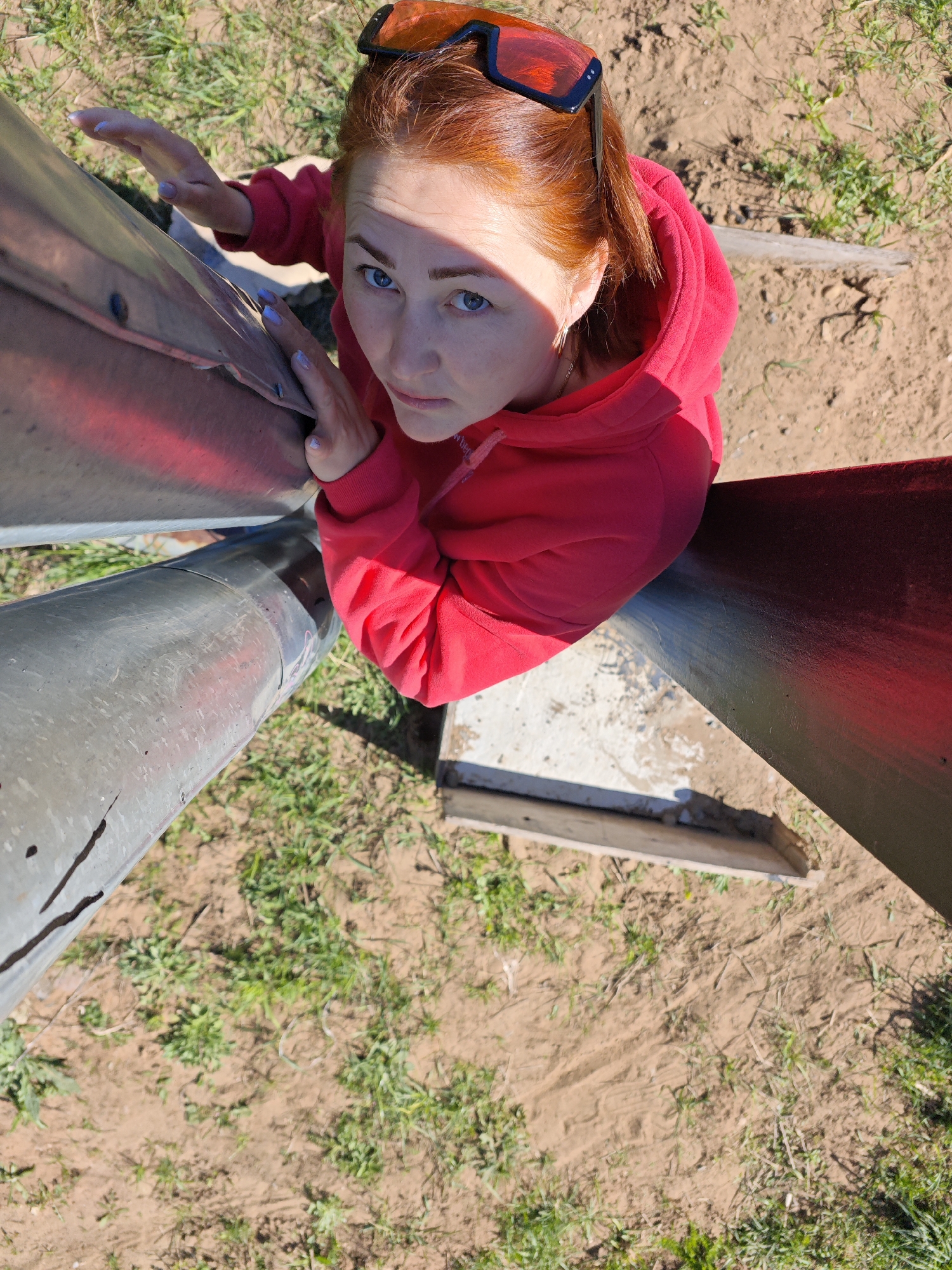 The image, taken from an extreme upward angle, features a red-haired woman wearing a red hoodie and sunglasses atop her head, situated amidst a sculptural element. The sculpture appears to consist of metallic, possibly repurposed, components. These elements include tubes and flat planes, some painted with red accents. The woman's hands are touching the metal structure. The ground is visible between the elements of the sculpture, exhibiting a mix of dirt and sparse vegetation.
Given the prompt, the location is assumed to be Марковский, Russia. However, the image itself does not provide any specific visual clues that would confirm this location.