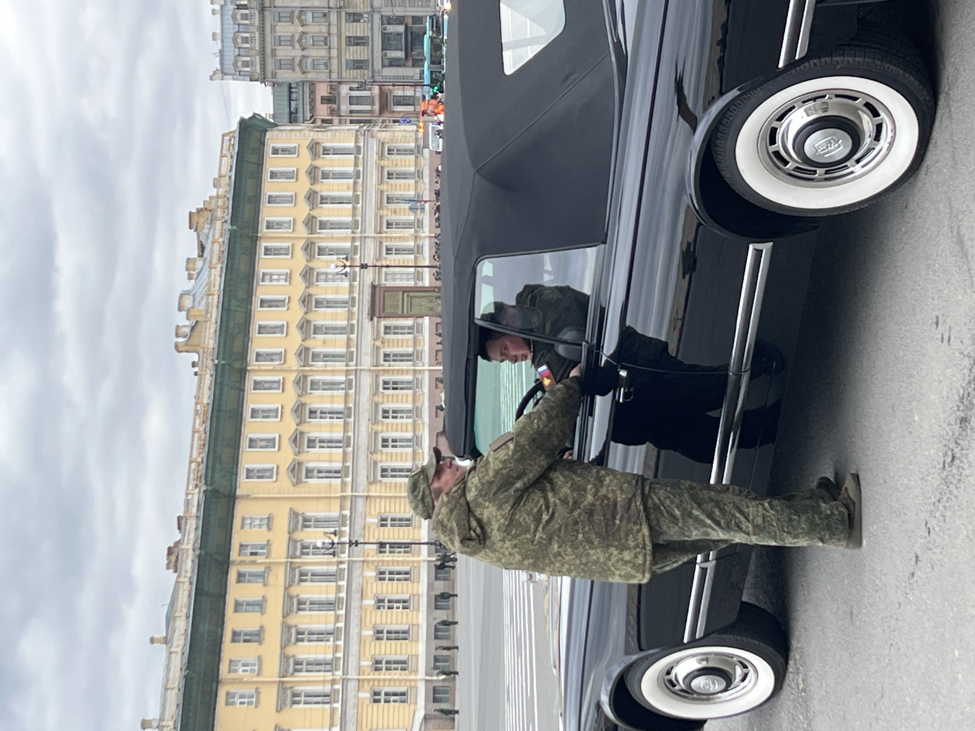 An outdoor, eye-level shot depicts a formal scene featuring a military figure and a classic car against a backdrop of historic buildings under an overcast sky.
In the foreground, a person in a modern camouflage military uniform, including a cap, stands beside a large, black, shiny convertible car. The individual appears to be leaning into the open driver's side window, possibly interacting with someone inside the vehicle or adjusting something. The car is sleek with chrome accents, and distinct whitewall tires with polished hubcaps featuring an unidentifiable emblem.
Reflected in the car's window and partially visible inside the vehicle is another individual, also appearing to be in military attire. A patch on their sleeve displays the colors of the Russian flag (white, blue, red horizontal stripes).
The background features stately, multi-story buildings, predominantly yellow with numerous windows, suggesting a European architectural style. One prominent building has a dark green roofline. Behind it, another, taller building with a more muted grey or brown facade is visible. The street appears paved and clear, implying an urban setting. The overall lighting suggests an overcast daytime, with soft, diffused light. There is no discernible specific text visible other than the potential flag colors. The scene conveys a sense of official engagement, possibly a parade, ceremony, or formal procession.