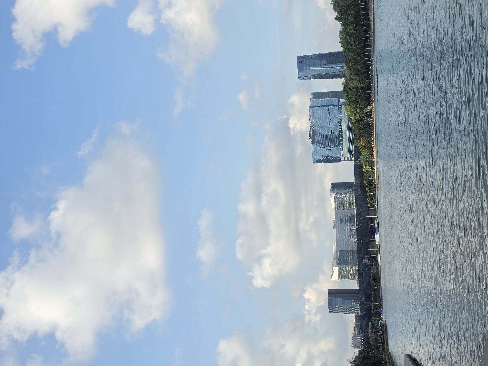 The image depicts a daytime view in Bangkok, Thailand, oriented with the sky on the left. A body of water, possibly a lake or river, fills the right side of the frame. Along the waterfront, modern buildings are visible, suggesting a business district. The presence of trees interspersed with the buildings indicates landscaping efforts within the urban setting. The sky is clear, with fluffy white clouds scattered throughout. The image is taken from an eye-level perspective, possibly from a waterfront promenade. No people are visible in the image.