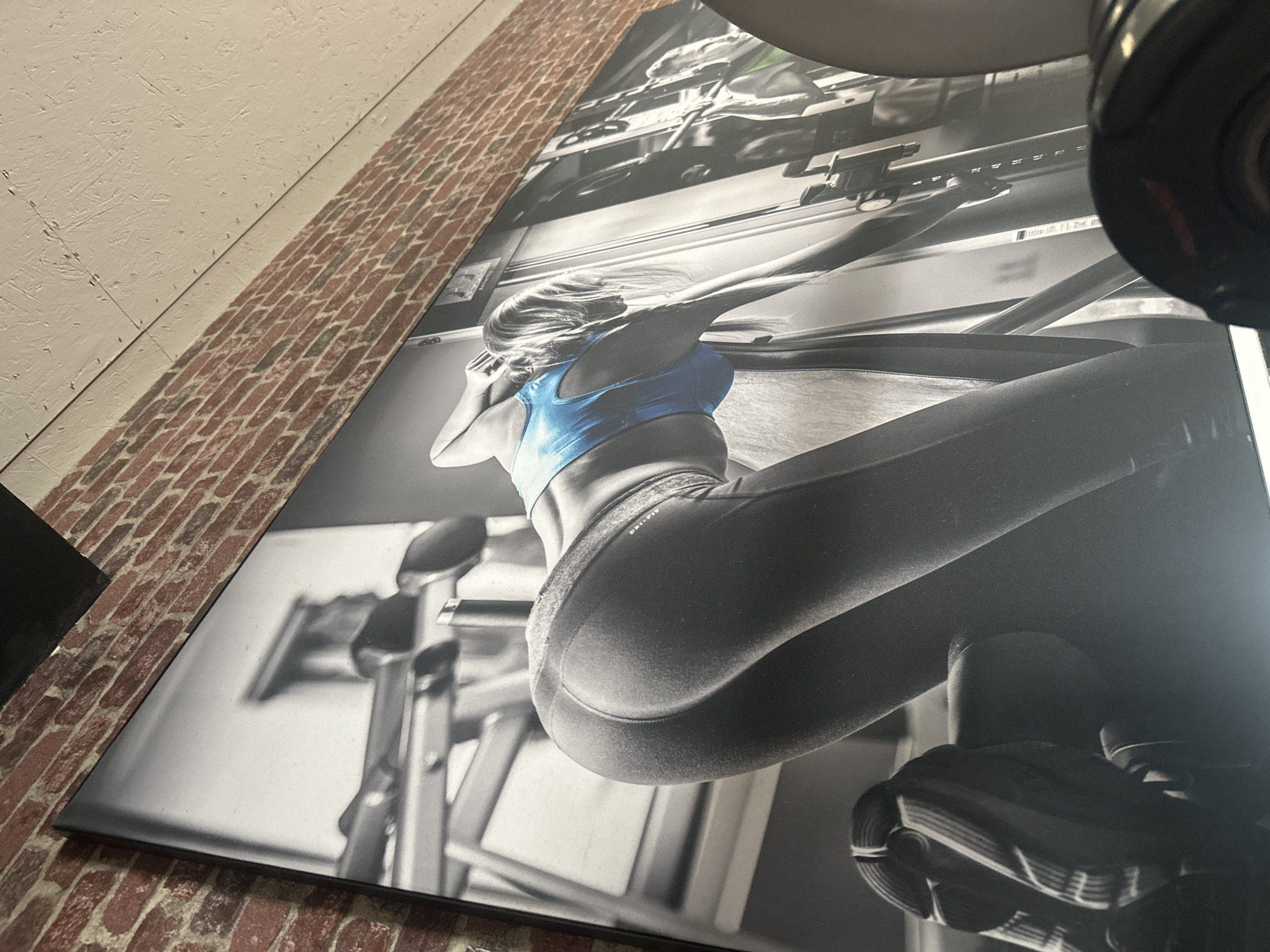 The image shows a poster of a woman using a Pilates reformer machine, mounted on a wall with brick veneer. The poster is black and white except for the woman's blue sports bra. The bottom corner of the poster sits on the brick veneer. The top right of the poster is obscured by a blurred object.
The location of the image is not ascertainable from the picture. There are no clues indicating the city of Brunnthal, Germany.