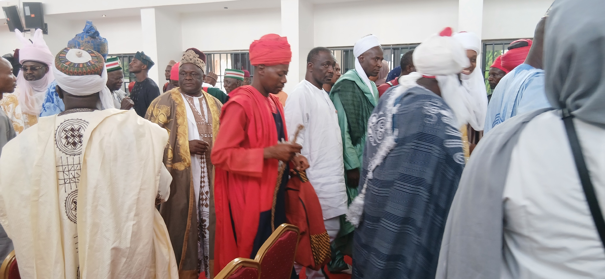 This image depicts a vibrant indoor gathering of numerous men in Ungwan Liman, Nigeria. The subjects are predominantly adult men, most of whom are adorned in traditional Northern Nigerian attire, including various styles of robes (such as babanriga or kaftan), elaborate turbans, and intricately embroidered caps (fila).
The scene suggests a formal or semi-formal social event or ceremony, with the men appearing to be moving and interacting within the space. In the foreground, with his back to the camera, a man wears a cream-colored robe featuring distinctive brown geometric embroidery on the back. He is also seen with a white turban topped by a decorated cap. Prominently featured in the center is a man in a striking red robe and matching red turban, holding what appears to be a ceremonial staff or whisk. To his left, another man wears a dignified brown robe embellished with rich gold embroidery on the trim and sleeves.
Other attendees are dressed in a variety of colors, including white, light blue, and dark green traditional garments. Many wear head coverings characteristic of the region. The setting is a well-lit indoor venue with light-colored walls, possibly a hall or a large room, indicated by modern ceiling lights and visible speakers in the upper left of one crop. A red upholstered chair is partially visible in the lower right corner. The expressions on the men's faces suggest engagement and participation in the ongoing event. There is no visible text in the image.