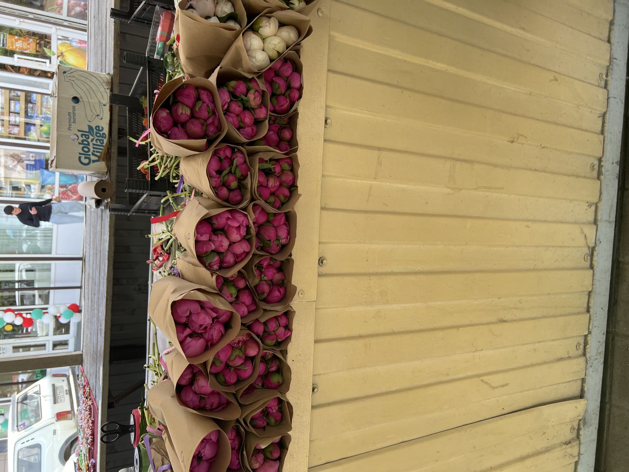 The image shows a street-side flower stall, presumably in Krasnodar, Russia. Bouquets of tightly-budded, mostly pink peonies wrapped in brown paper are displayed in a vertical arrangement. A few bouquets of white flowers are visible at the top. Next to the flowers is a yellow, corrugated metal wall, which forms part of the stall's structure. In the background are street scenes with a man in a blue jacket, storefronts, and a white van. A box labeled "Global Village" is also present. The presence of flowers, and street-side activity indicates a commercial area. The presence of the "Global Village" box gives the possibility that international products are sold in this area.