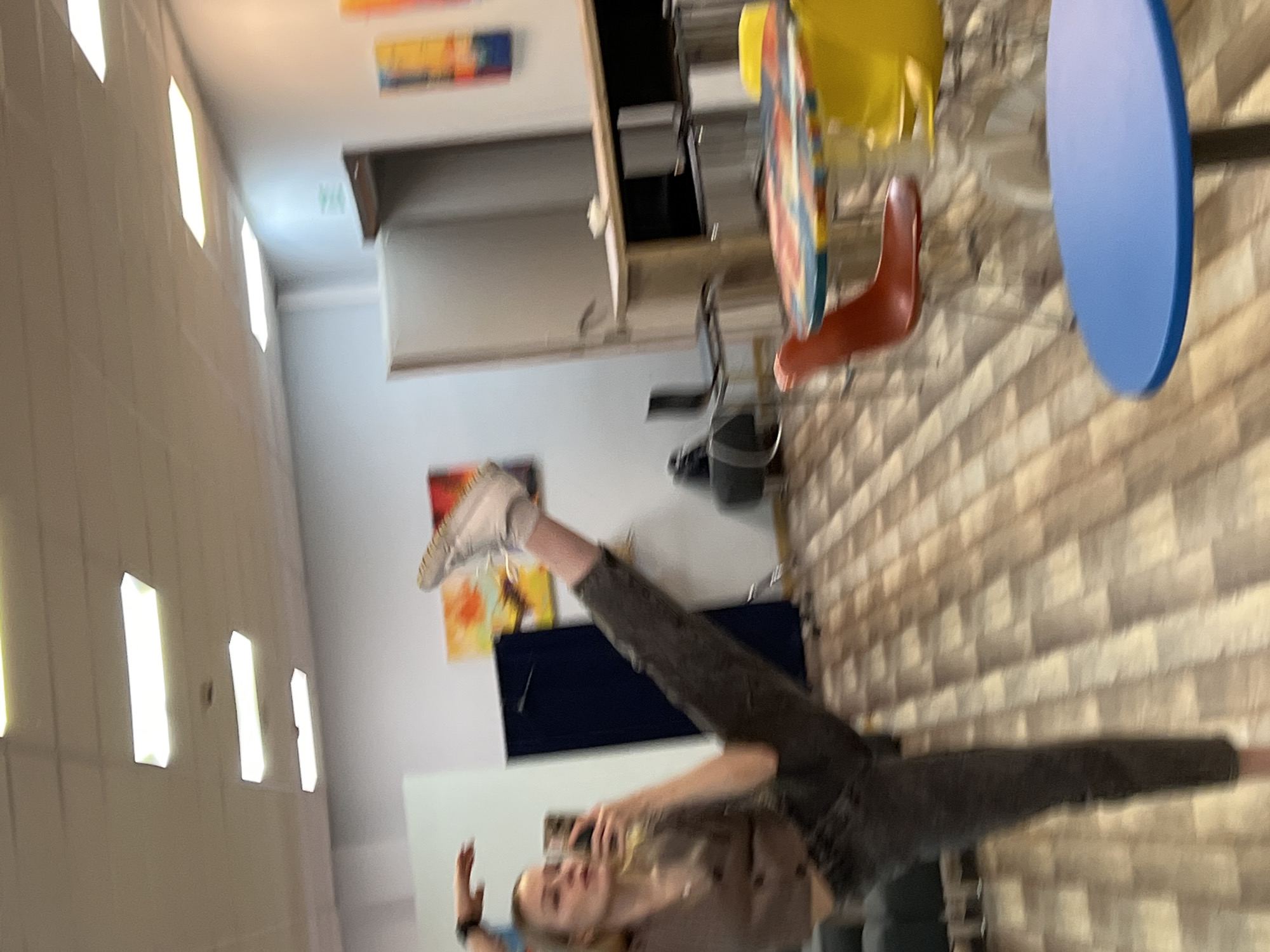 A person is performing a leg split indoors. They are holding a smartphone, presumably taking a photo of themself. The background includes a dining area with tables and chairs, kitchen countertops, artwork on the walls, and artificial lighting. The location is Khimki, Russia.