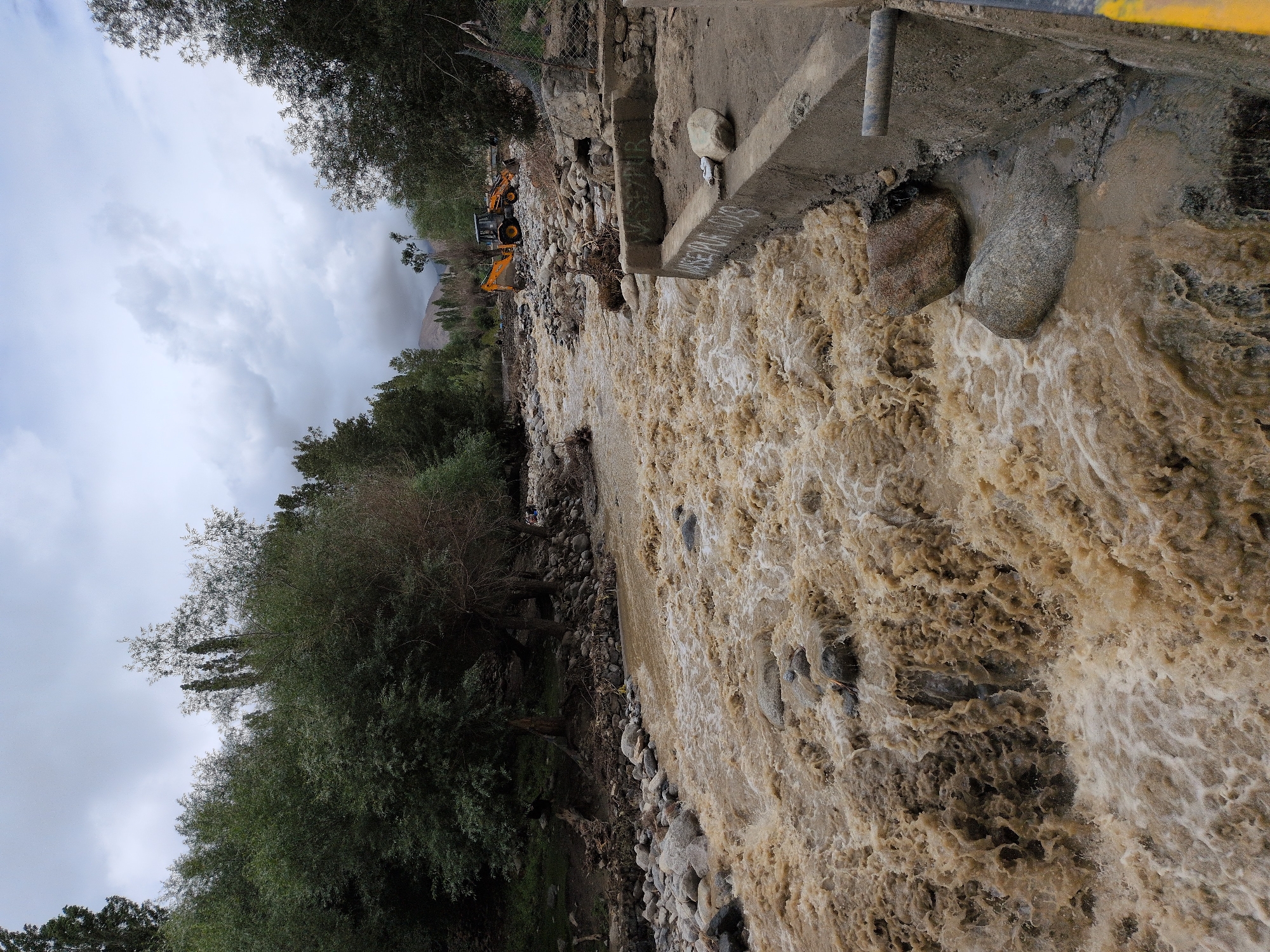 The image depicts a turbulent, muddy river flowing through a rocky gorge, likely in Leh, India, given the mountainous terrain and vegetation. The water is a rich, opaque brown, indicating a high sediment load, and churns with white foam as it rushes downstream. Large, grey rocks are scattered along the riverbed and banks, some partially submerged.
On the right bank, a concrete structure, possibly a bridge or retaining wall, shows signs of damage and debris. Graffiti is visible on the concrete. Adjacent to the river, a yellow backhoe loader is parked, suggesting ongoing or recent work, potentially related to flood control or cleanup. Trees with green foliage line the left bank and the upper reaches of the gorge, creating a contrast with the barren, rocky slopes and the muddied water. The sky is overcast with heavy grey clouds, contributing to the somber mood of the scene. A wire mesh fence runs along the right bank in the distance. There are no people clearly visible in the immediate foreground or mid-ground, though faint figures may be present further upstream on the left bank. The overall impression is one of a natural force, a flood, impacting the landscape and requiring human intervention.