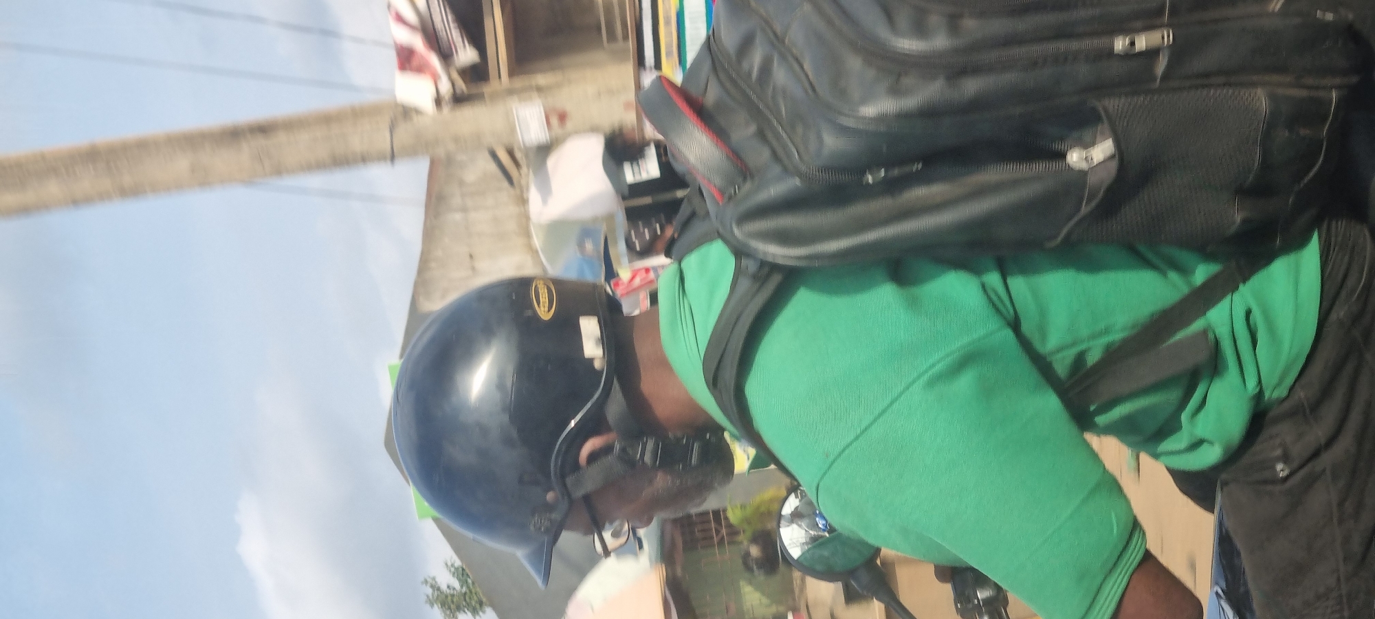 An individual wearing a black helmet and a green shirt rides a motorcycle, carrying a black backpack. The scene takes place outdoors in Otta, Nigeria, with buildings and a utility pole visible in the background. The sky is partly cloudy. The individual's face is partially obscured by the helmet and glasses, and their left arm is resting on the motorcycle handlebar. A rearview mirror is attached to the motorcycle.