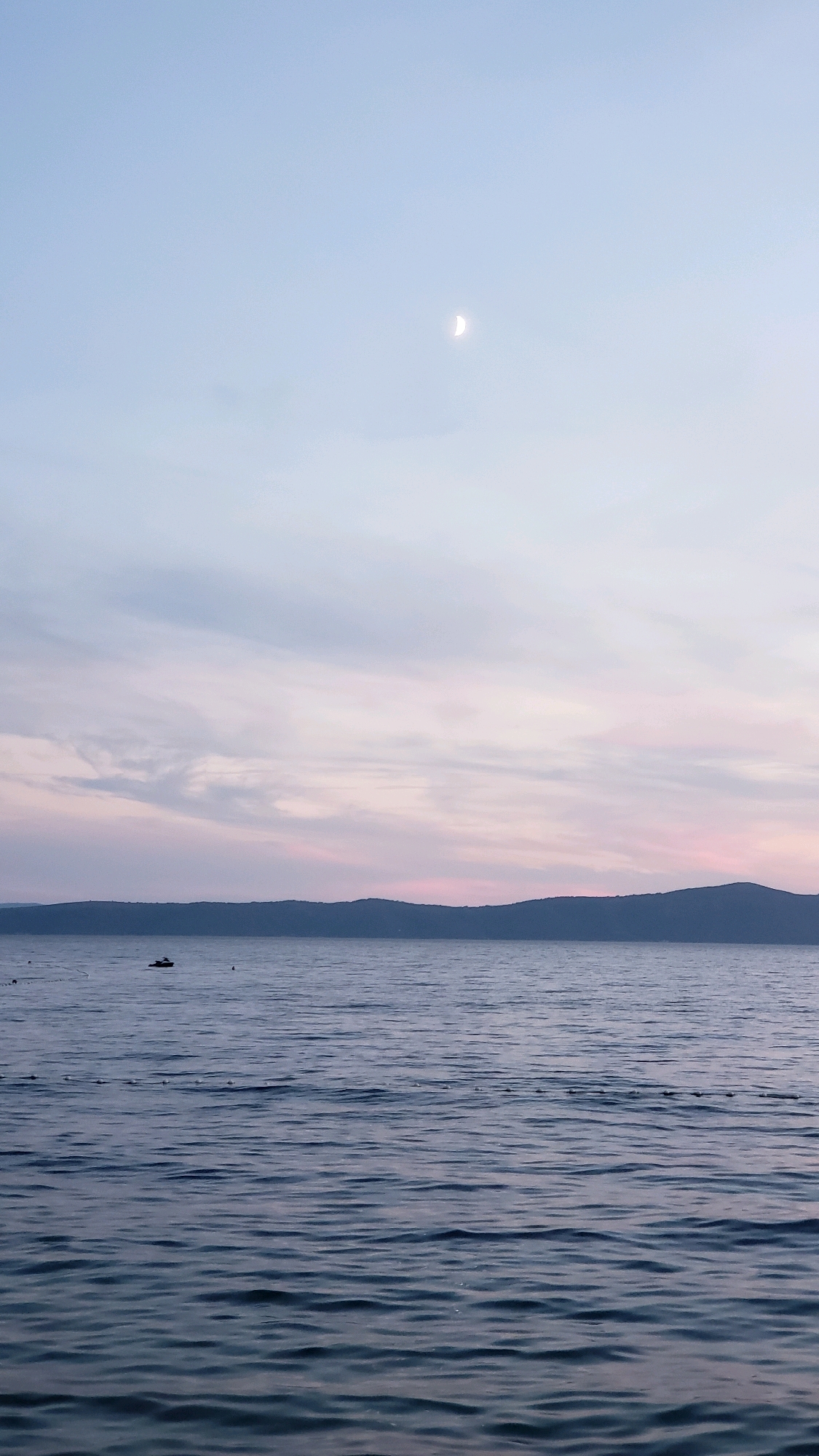The image depicts a serene seascape at dusk, likely in Općina Tučepi, Croatia, given the subtle hint of the Adriatic Sea. The sky is a soft gradient of pale blue at the top, transitioning to delicate hues of pink and lavender as it meets the horizon. A crescent moon is visible in the upper portion of the sky. Below the sky, a dark, undulating silhouette of hills forms the distant coastline. The middle ground is dominated by the calm surface of the sea, reflecting the muted colors of the sky. A small, dark-colored boat is positioned on the water, approximately one-third of the way from the left edge of the frame and close to the horizon line. A series of small, dark, rounded objects are arranged in a line on the water's surface in the lower left portion of the image, possibly buoys or markers. The foreground consists of more of the tranquil sea, with gentle, textured ripples creating a sense of depth and movement. The overall atmosphere is peaceful and tranquil, characteristic of a calm evening by the coast.