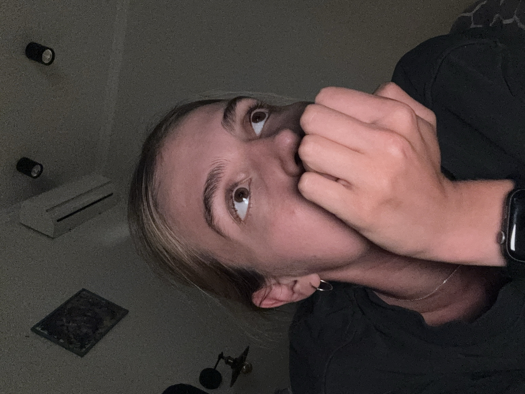 A close-up, rotated image features a fair-skinned individual with light brown eyes and blonde hair. Their face is partially obscured by their own hand, positioned near their mouth. They are wearing a dark-colored shirt and a thin silver necklace. An Apple Watch is visible on their wrist. The background includes two cylindrical light fixtures on a wall, an air conditioning unit, a small framed picture on the floor, and a microphone stand. The location is Unknown, Unknown.