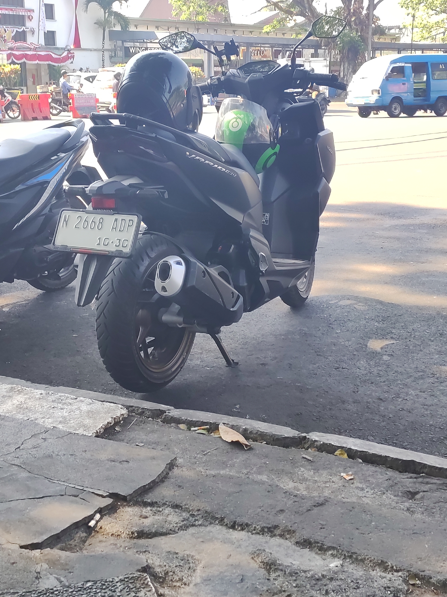 The image captures a street scene in Malang, Indonesia, focusing on a parked black scooter in the foreground. The scooter's license plate is clearly visible, reading "N 2668 ADP" with additional text below it. A black helmet rests on the back of the scooter, and a green Gojek helmet is visible in the front compartment. To the left of the scooter, another partially visible scooter is parked.
In the background, a blue van is in motion on the street, and there are buildings and trees that suggest an urban environment. Pedestrians are present in the background, with one individual appearing to be a delivery person. The street is paved, and the sidewalk in the foreground shows signs of wear and tear, with cracked concrete. The lighting suggests it is daytime, with shadows cast by the sun. The scene appears to be a typical urban setting in Malang.