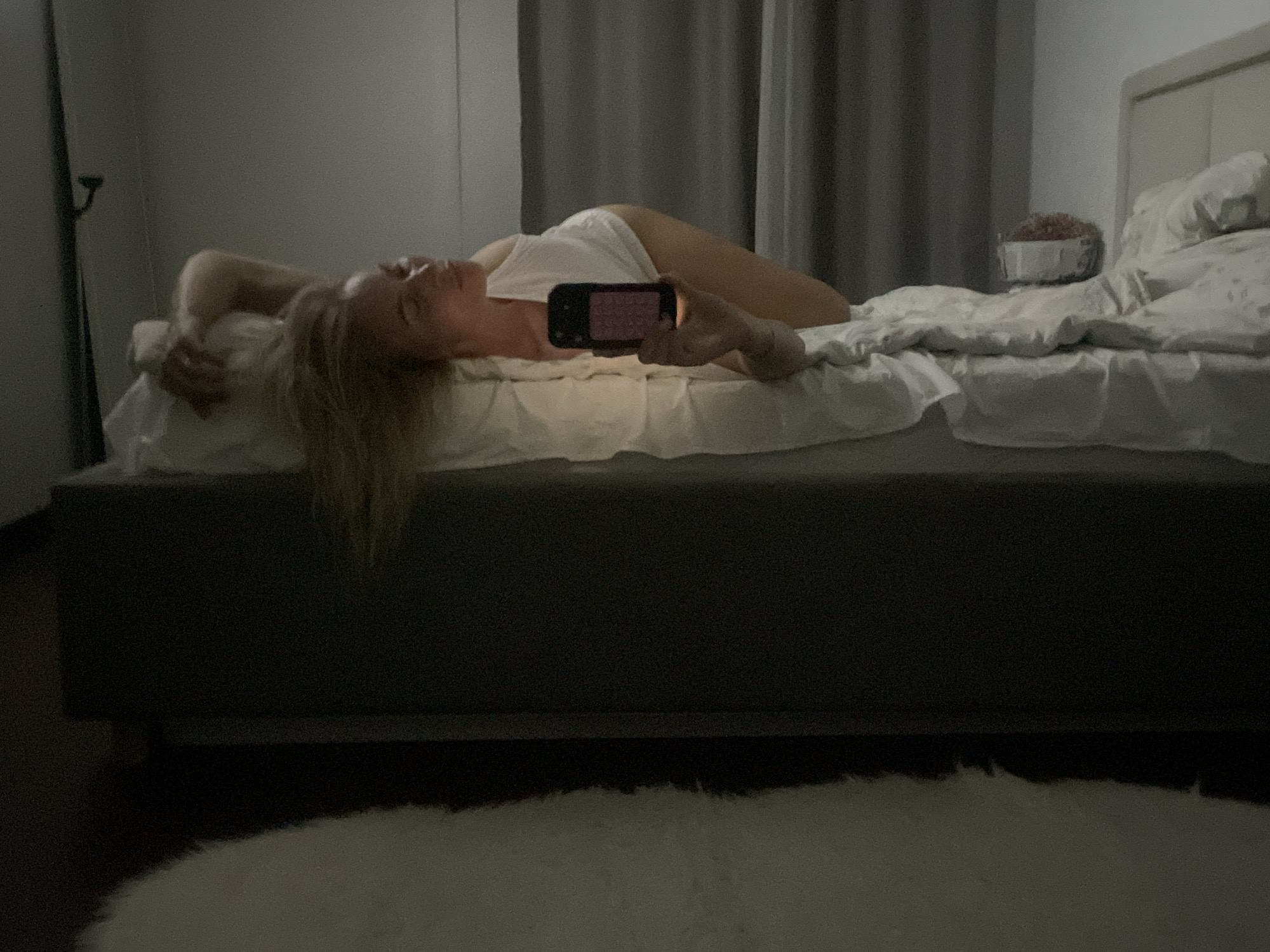 The image shows a person lying on a bed. The person is holding a phone, potentially taking a selfie. The bed has white linens and a dark-colored frame. A small table with items sits next to the headboard. A rug is on the floor. The environment appears to be a bedroom. There is no information to locate the scene in Baranovka, Russia.