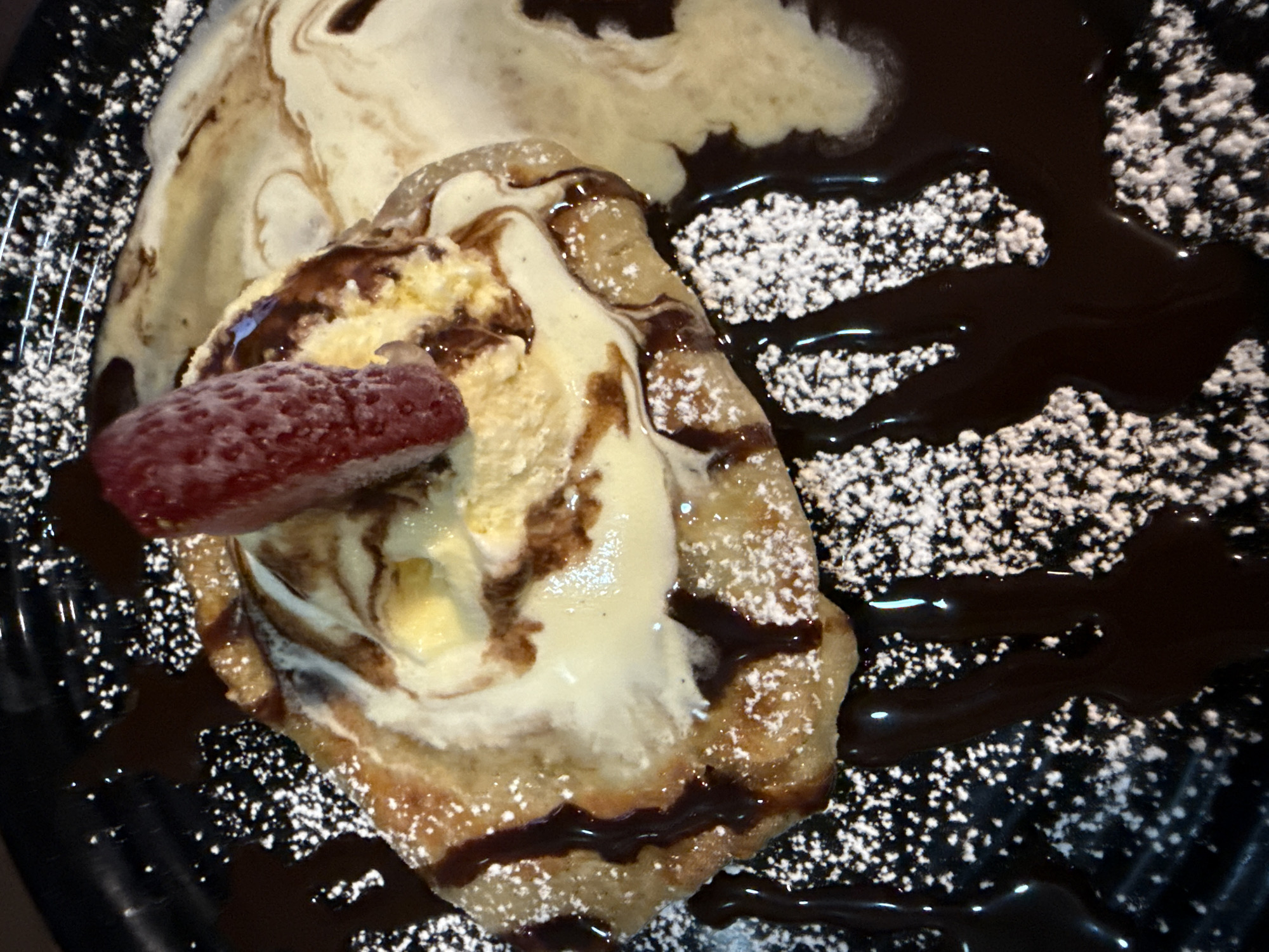 A dessert featuring a pastry, likely a crepe or similar item, topped with ice cream, chocolate sauce, powdered sugar, and a strawberry, is presented on a black plate. The plate also exhibits a large swirl of cream or icing. The scene suggests a culinary setting, possibly a restaurant or cafe in Puebla, Mexico, given the prompt. No people are visible.