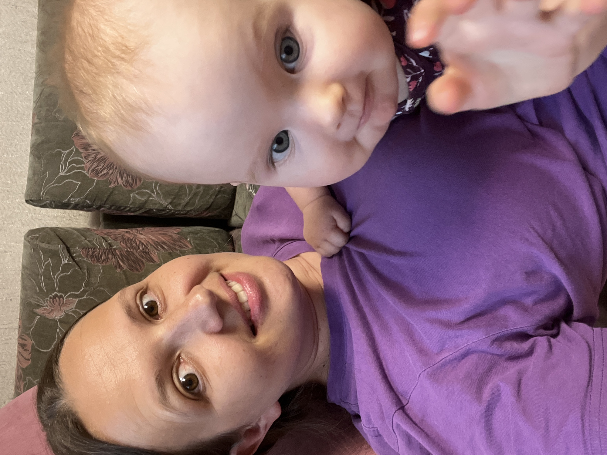 A selfie-style image shows a woman and a baby on a couch. The woman is in the foreground on the left, looking at the camera with a smile. She has brown hair and brown eyes and is wearing a purple t-shirt. The baby is on the right, leaning against the woman, with its face close to the camera. The baby has blonde hair and blue eyes and is looking slightly to the side. The baby's right hand is raised towards the camera. The couch has a dark green fabric with a floral pattern. The background is slightly blurred, showing the couch and a wall. The image appears to have been taken indoors.