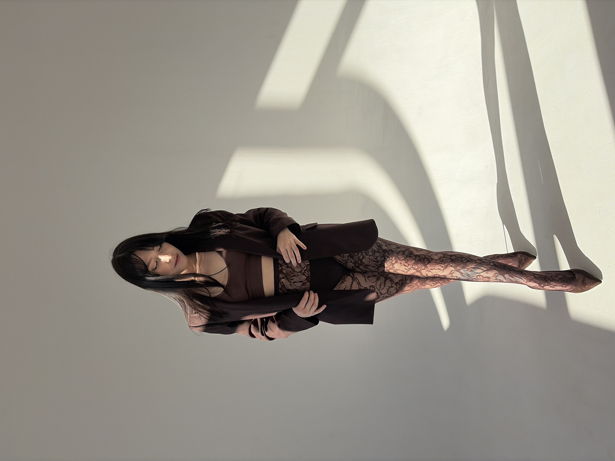 A woman with long dark hair is lying on a light gray surface, possibly a floor or large table, in a brightly lit area. She is wearing a brown crop top, dark brown shorts, and a matching brown blazer. She also has patterned tights on her legs. The light appears to be coming from a window or skylight above, creating sharp, elongated shadows on the surface next to her. The location is unknown as is there any verifiable evidence this is in Chaikovsky, Russia.