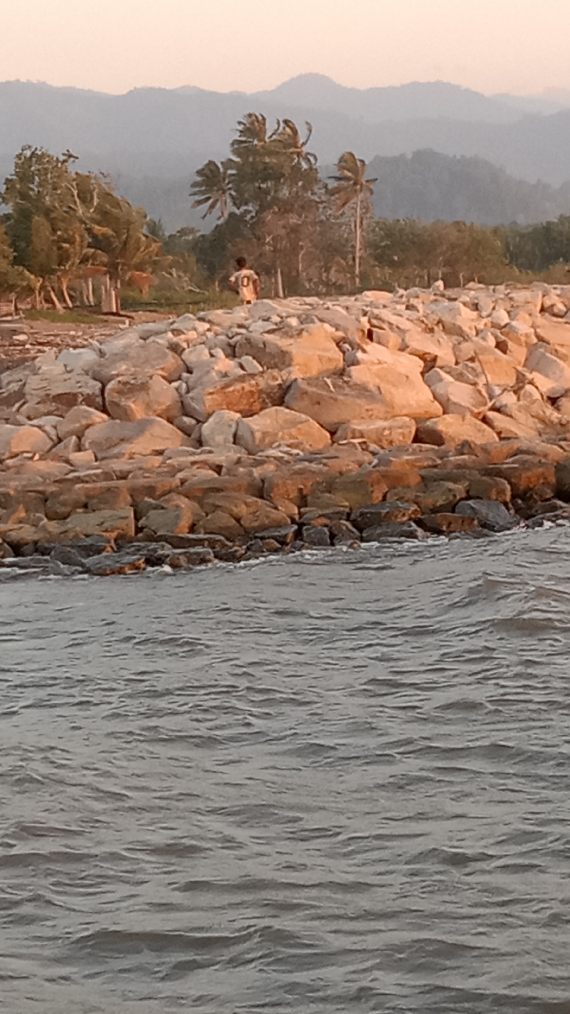 The image depicts a person walking on a rocky shoreline. The person is wearing a light-colored t-shirt with a dark number "10" on the back. They are positioned towards the center of the frame, facing away from the viewer and towards a line of trees and hills in the distance. The shoreline is made of large, rough-hewn rocks, with smaller rocks and debris scattered around. The water in the foreground is choppy, suggesting wind or a current. In the background, a range of hazy, tree-covered hills forms the horizon, silhouetted against a pale, dusky sky. The scene is set in an outdoor, natural environment, likely a coastal area in Indonesia, given the presence of palm trees. The overall impression is one of solitude and a serene, natural landscape at dusk.