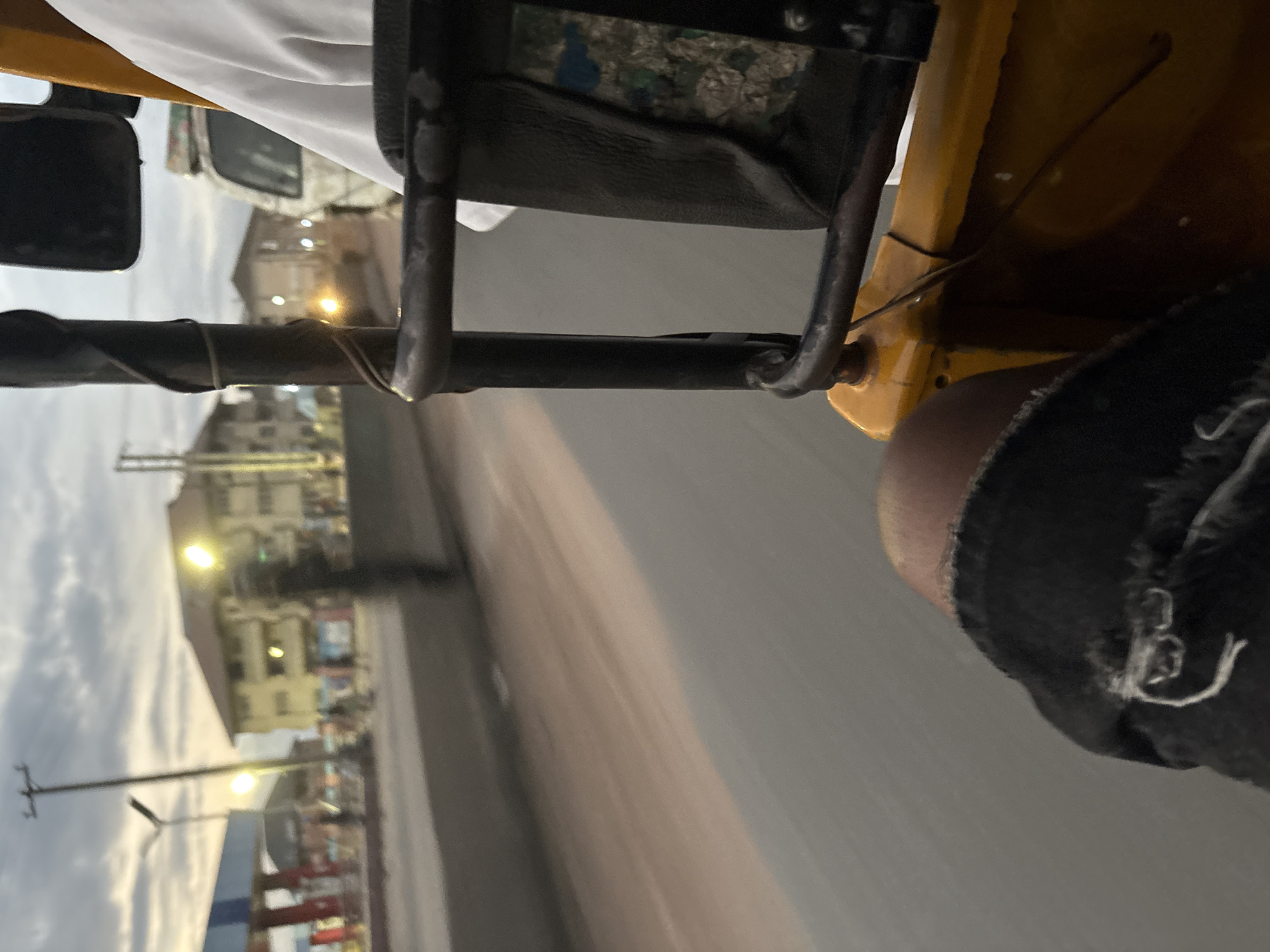 An outdoor, eye-level, medium shot shows a first-person perspective of someone riding in the passenger seat of a yellow auto-rickshaw in Onitsha, Nigeria, during dusk. The passenger is wearing ripped black jeans. The auto-rickshaw is moving, as evidenced by the motion blur of the road and the blurred buildings in the background. A person in a white shirt is visible in the passenger seat next to the camera. A white van is seen in the distance, also in motion. The sky is cloudy and overcast, indicating the end of the day. A gas station with a blue and white canopy is on the left side of the image, and several buildings are visible across the street. The street appears to be a busy thoroughfare, with several blurred figures walking along the sidewalk. The overall atmosphere is one of a typical urban scene in Nigeria at twilight.