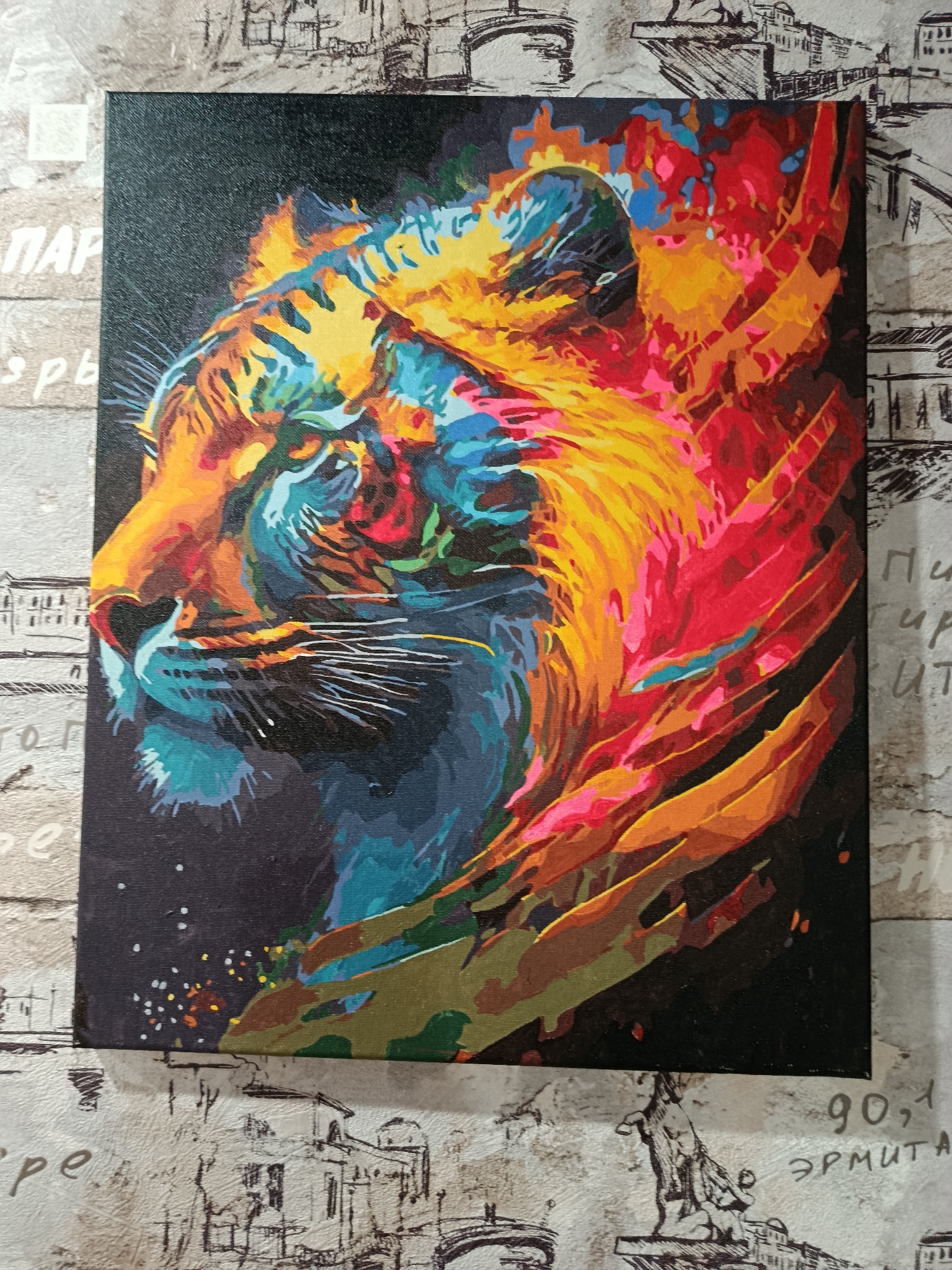 The image is a close-up, slightly angled, top-down view of a painting of a tiger's head, rendered in a vibrant, abstract style with bold strokes of color. The tiger's face is depicted in profile, facing left, with intense blues, greens, yellows, oranges, and reds dominating its features. The background of the painting is black, with small white and colored dots scattered across it, resembling stars or splatters.
The painting is placed on a surface with a patterned wallpaper. The wallpaper has a light, textured background with a repeated print of various European architectural elements, possibly a cityscape or historical buildings, rendered in thin, dark lines. There are also some indecipherable handwritten words and numbers printed on the wallpaper.
The photograph does not provide enough information to determine its location in the city of Apatity, Russia.