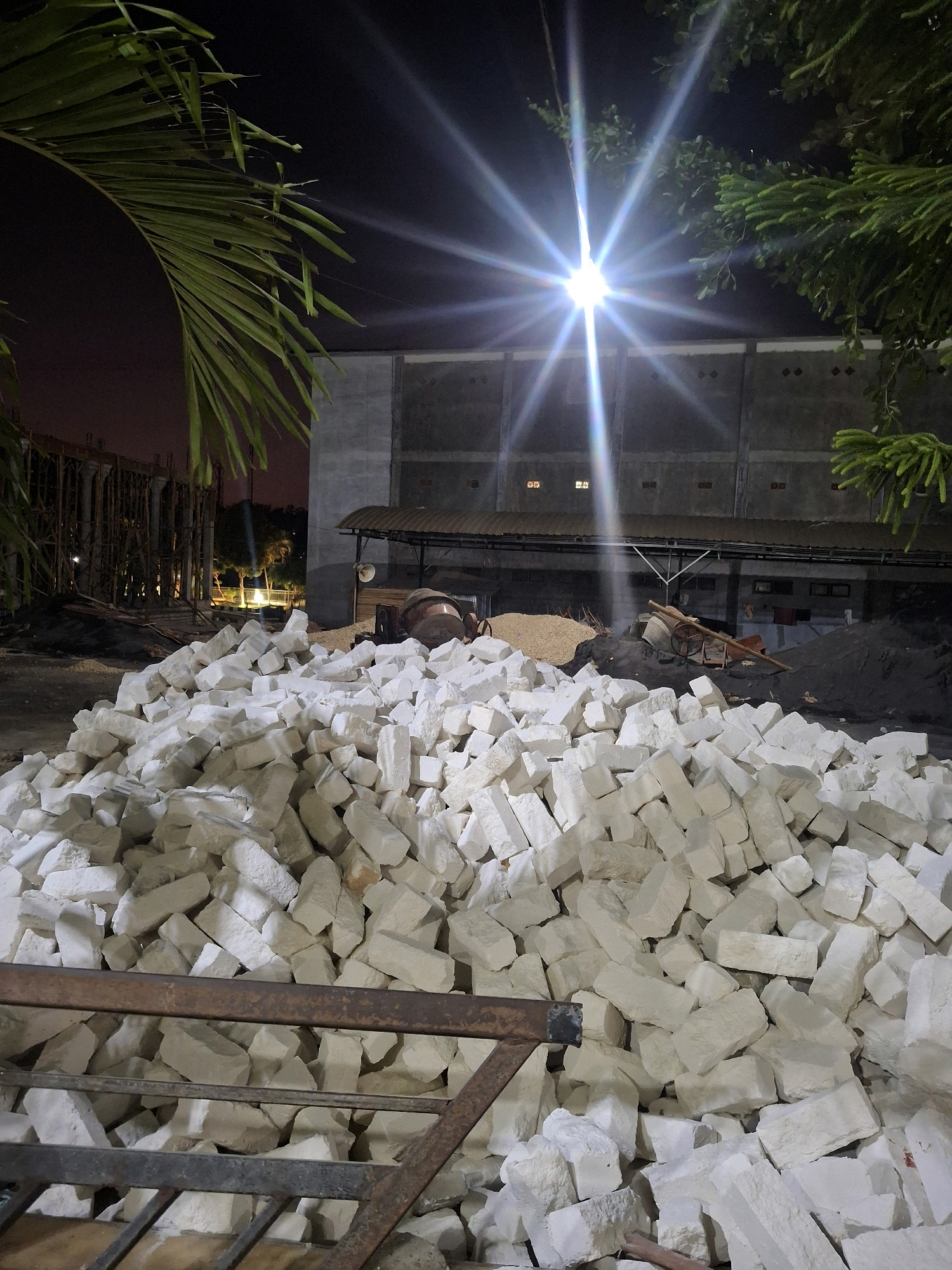 The image depicts a nighttime scene at a construction site in Potoan Laok, Indonesia. In the foreground, a large pile of white bricks is visible. Behind the bricks, a cement mixer and a small cart are present. The background shows a building under construction, with scaffolding and structural elements. A bright light source, possibly a street lamp, illuminates the scene, creating a starburst effect. Trees and foliage frame the right side of the image. The overall impression is of a work in progress during nighttime hours.