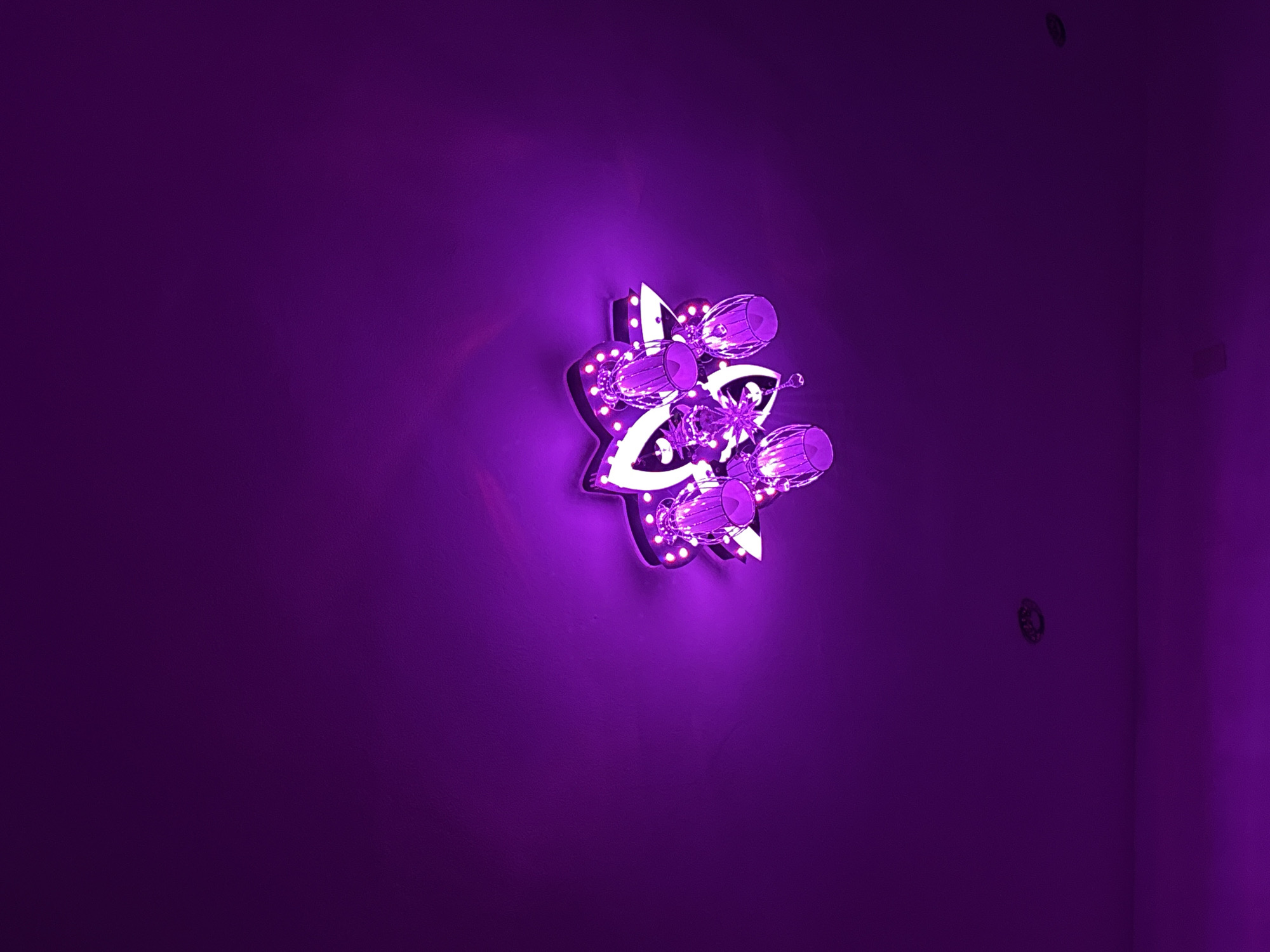A vibrant purple wall is illuminated by a decorative, star-shaped light fixture centrally mounted. The fixture, characterized by a geometric design, features multiple small, round LED lights embedded along its edges, emitting a bright white-purple glow. It is adorned with clear, crystalline embellishments, including several cylindrical elements and smaller star-shaped accents that refract the light.
The intense illumination from the fixture creates a radiant purple halo on the immediate wall surface, fading into a deeper purple further away. The wall itself exhibits a subtle, granular texture. In the upper right corner of the frame, a small, dark, circular object, possibly a screw head or wall plug, is visible. Towards the lower right, a dark vertical line suggests a corner of a room or an adjacent wall, where a faint, small, dark rectangular object, possibly an electrical outlet or fixture, is present. The scene is indoors, presenting a distinct and modern aesthetic dominated by shades of purple. The context places this scene in Gorno Sahrane, Bulgaria. No text is visible in the image.