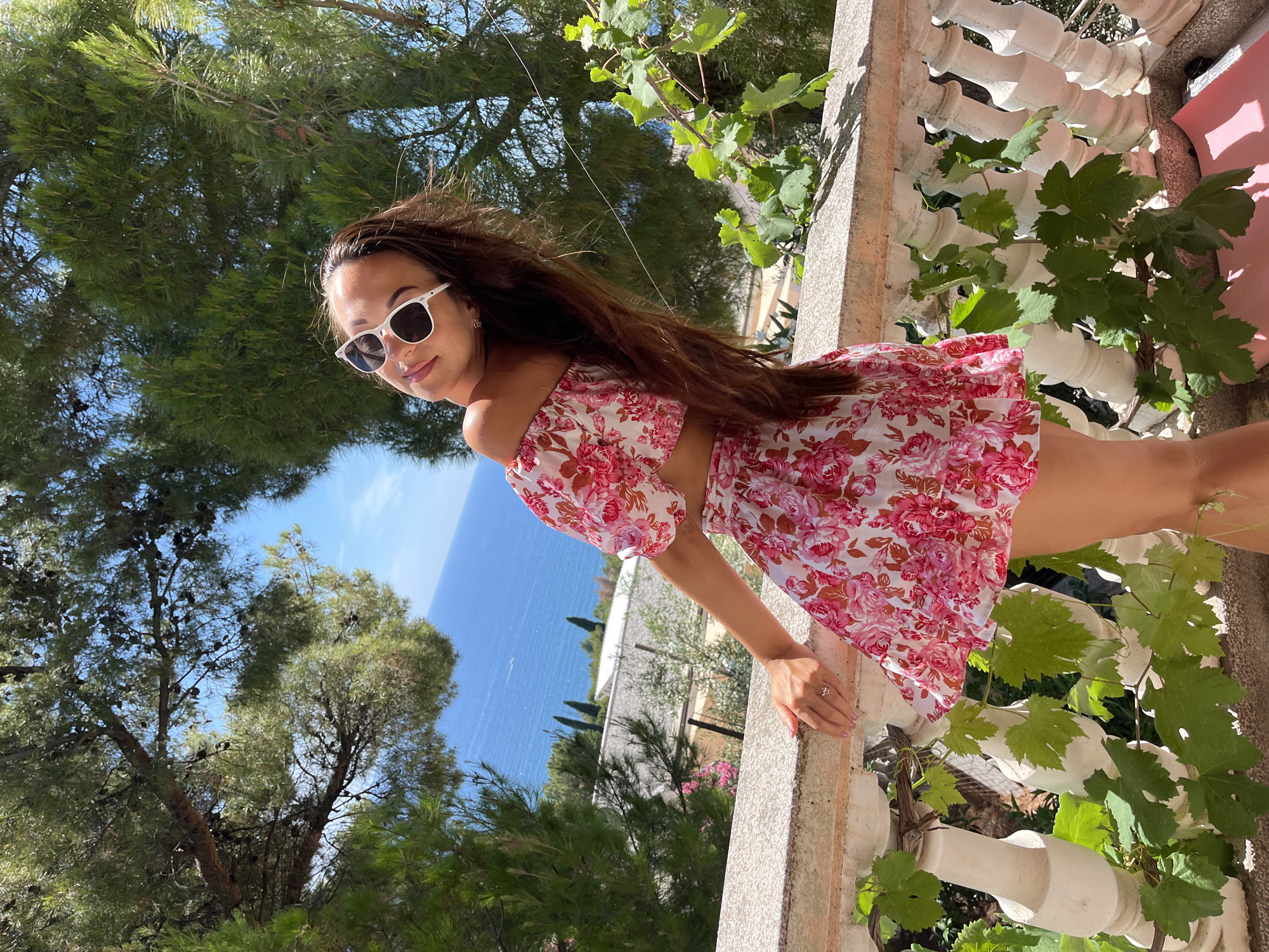 A young woman stands on a balcony overlooking the sea in Šušanj, Montenegro. She is wearing a pink floral off-the-shoulder crop top and a matching skirt, white sunglasses, and a ring on her left hand. Her long, brown hair is flowing to her right. The balcony is made of stone and features a white railing with ornate balusters, some of which are covered in green vines. In the background, the Adriatic Sea is visible, with a clear blue sky above. There are trees with pinecones and foliage surrounding the balcony. The overall impression is one of a sunny, vacation-like setting.