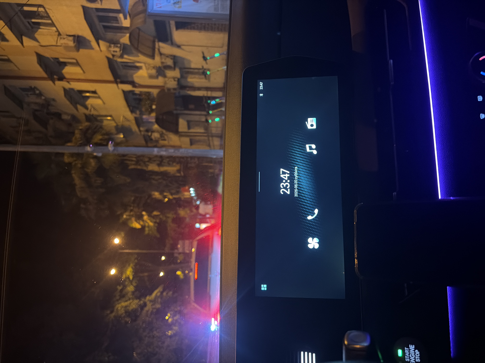The image shows the interior of a car at night, looking out through the windshield and at the car's dashboard display. The dashboard shows the time as 23:47 and the date as 2026-06-20. Icons for a fan, a phone, music, and radio are visible on the screen. Outside the car, streetlights illuminate a street with buildings and palm trees on the right. There are several electric scooters parked along the sidewalk. The car's interior has blue ambient lighting. The overall scene suggests a residential area at night.