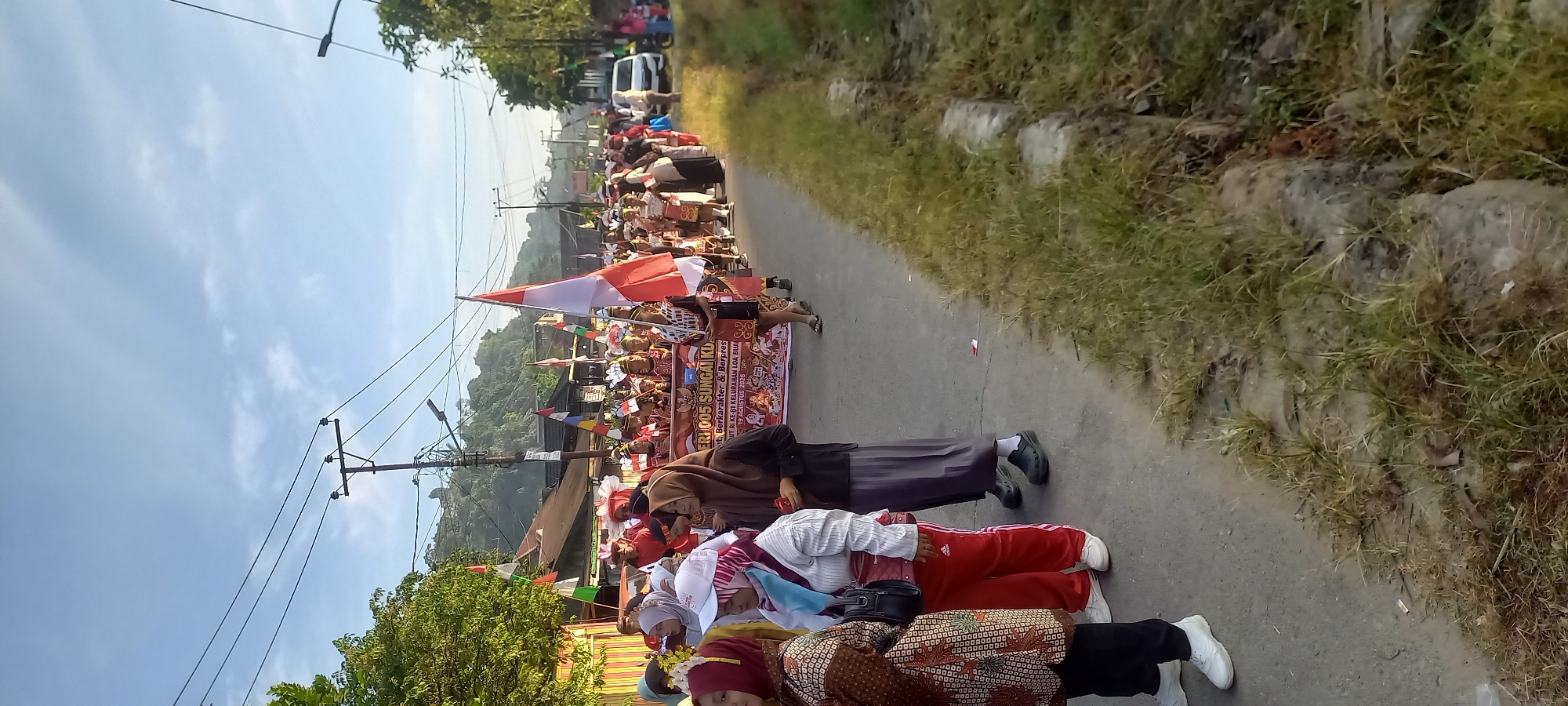 The image depicts a street scene in Samarinda, Indonesia, likely during a parade or celebration. In the foreground, several individuals are walking on a paved road. To the left, a woman in a patterned brown top and black pants, wearing white sneakers, is visible. Next to her, a woman in a white shirt, red pants, and white sneakers is also walking. Further back, a woman in a brown hijab and grey skirt is seen, wearing black clogs.
The middle ground of the image shows a procession of people, many wearing traditional attire and headdresses. A large banner is being carried by a group of individuals. The banner has text in Indonesian, which appears to include "KE-81 KELURAHAN LOA BULAN" and a date "17 AGUSTUS 2026". Several Indonesian flags (red and white) are being waved and carried by participants. Other flags with various colors are also present. The procession is moving along the right side of the road.
In the background, there are buildings lining the street, some appearing to be shops or houses. A hill with trees is visible behind the buildings. Overhead, power lines and utility poles are present, crisscrossing the sky, which is partly cloudy. A white car is parked on the right side of the road. The environment appears to be an urban or semi-urban area. The lighting suggests it is daytime, possibly morning or late afternoon due to the low angle of the sun casting long shadows. The overall atmosphere is festive and communal.