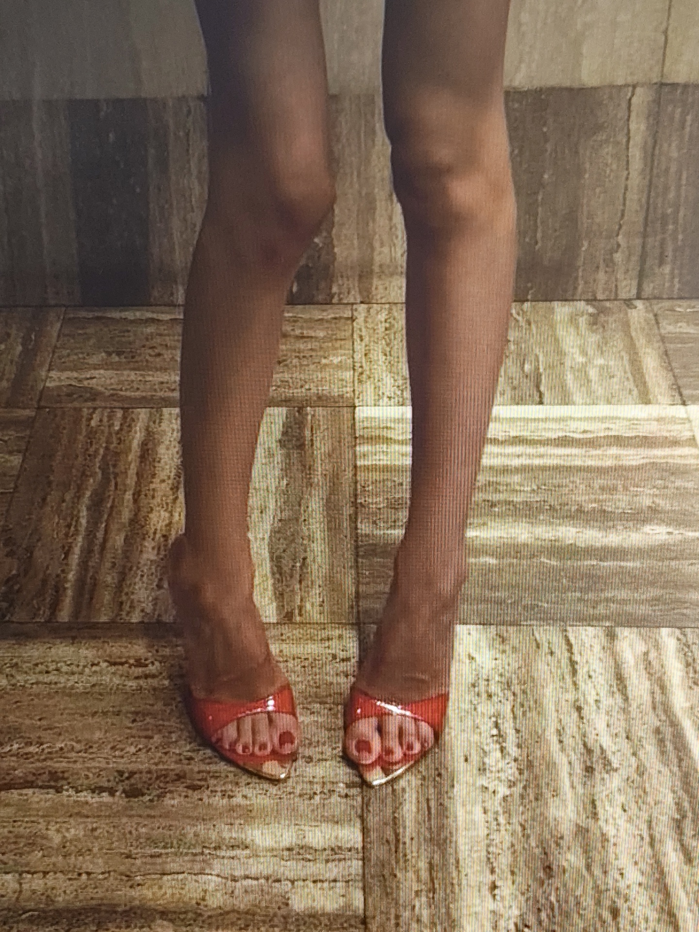The image shows a close-up view of a person's legs and feet, wearing bright red, open-toed high heels. The person is standing on a floor with a patterned surface, which appears to be made of tiles with a marble-like texture. The background is out of focus but also seems to be a wall with a similar textured finish. There are no discernible elements in the image that would allow for identification of the location within the city of Il'inskiy, Russia. The lighting suggests an indoor setting.
