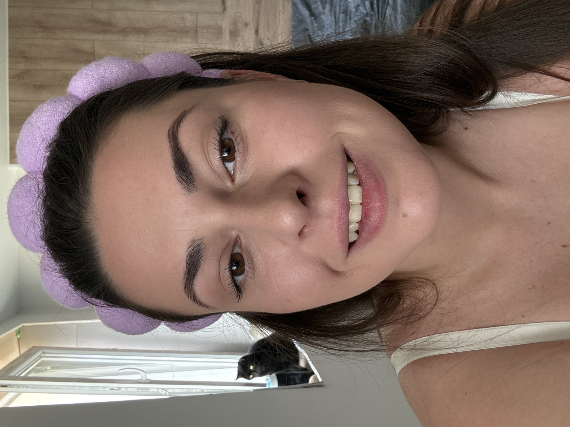 A close-up, selfie-style shot shows a woman with dark hair and a lavender headband. She is wearing a white tank top and has makeup on. Her mouth is slightly open in a smile, revealing her teeth. In the background, a black cat is visible through a window to the left, sitting on a ledge. To the right, a bed with dark grey bedding is partially visible. The overall environment appears to be an indoor setting, possibly a bedroom.