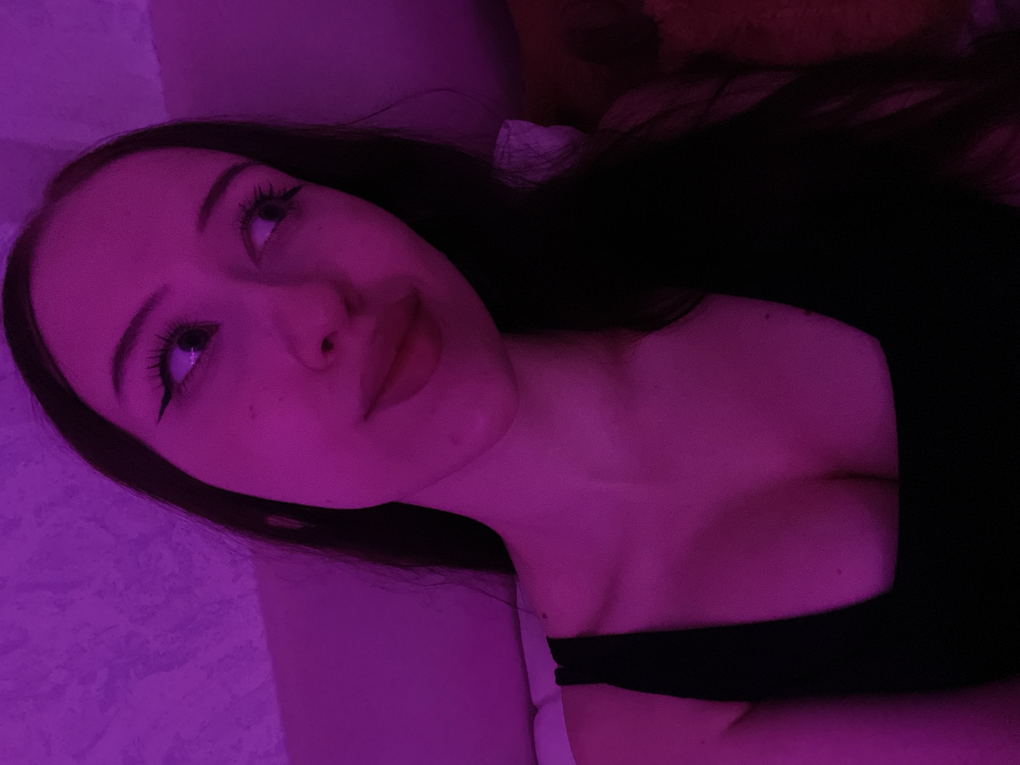 A young woman with long dark hair and full lips is captured in a close-up, medium shot, illuminated by purple lighting. She wears a black tank top that reveals her décolletage. Her eyes, with dramatic winged eyeliner, gaze upwards and to the right. The background is a textured wall, also bathed in purple light. The scene appears to be indoors, with a soft surface visible behind her left shoulder. There are no discernible landmarks or environmental cues that would indicate a specific location within Votkinsk, Russia.