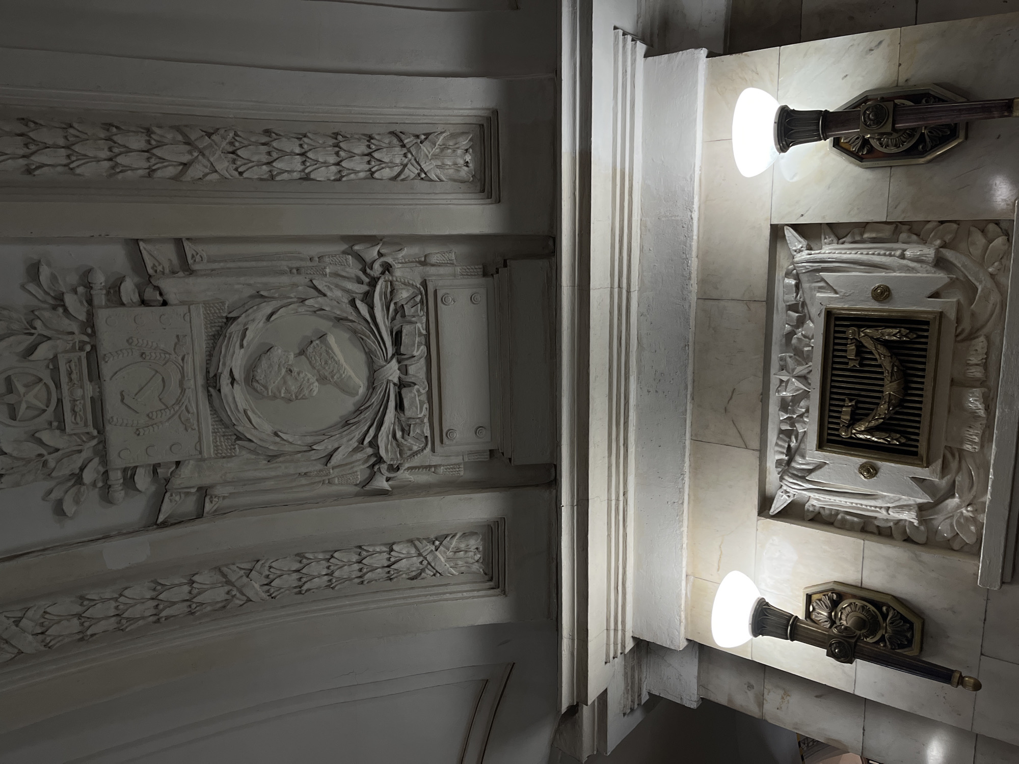 This is a low-angle, medium shot of decorative architecture in the Moscow Metro. The image focuses on ornate plasterwork on the arched ceiling and the upper section of a wall.
The ceiling features repeating patterns of stylized leaves and bands of relief ornamentation. Centered on the ceiling is a prominent relief panel depicting a star, a hammer and sickle, and wheat stalks, signifying Soviet iconography. Below this, a circular bas-relief profile of a man in a military-style uniform is framed by laurel wreaths.
The wall below the ceiling is faced with marble. Two ornate sconce lights, resembling torches, are mounted on the marble. They have dark bases with decorative bronze or brass elements and emit a bright, diffused light from white frosted glass shades.
Between the sconces, a large, decorative plaster frieze is mounted on the marble wall. This frieze incorporates a central grille-like element, flanked by draped fabric motifs and stars. The overall aesthetic is grand and monumental, characteristic of Stalinist architecture found in many Moscow Metro stations. The scene is devoid of people, focusing solely on the architectural details and lighting.