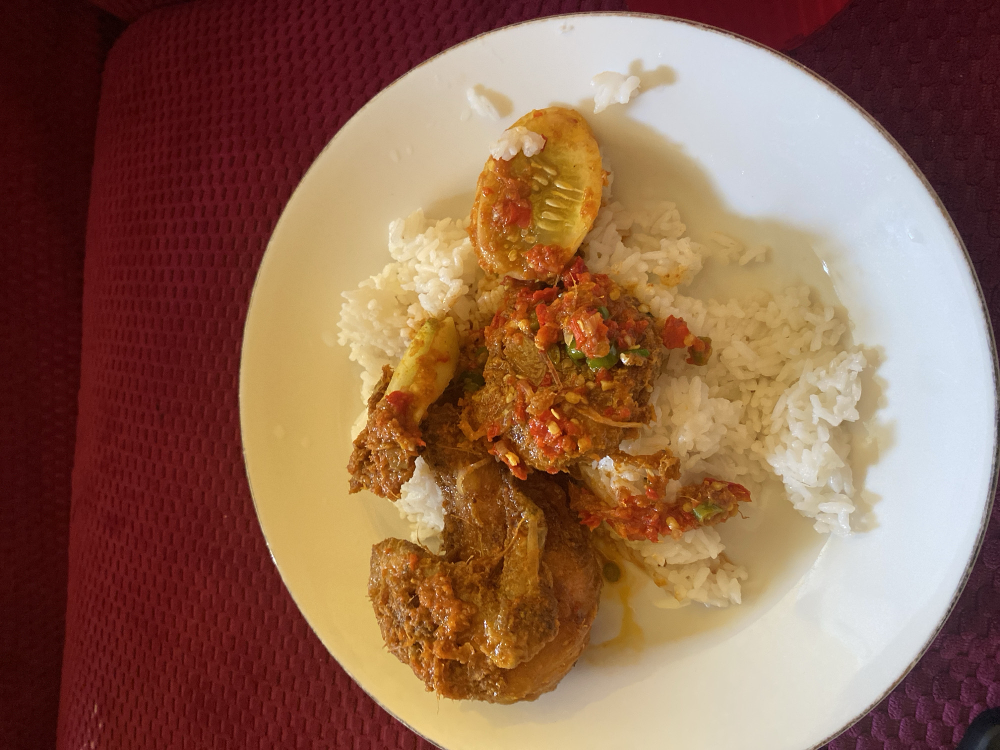 A white plate with a brown rim contains a meal of white rice, chicken pieces in a reddish-brown sauce, and sliced cucumber. The plate is set on a textured dark red surface, possibly upholstery. The lighting is overhead, casting soft shadows. The scene appears to be in a domestic setting.