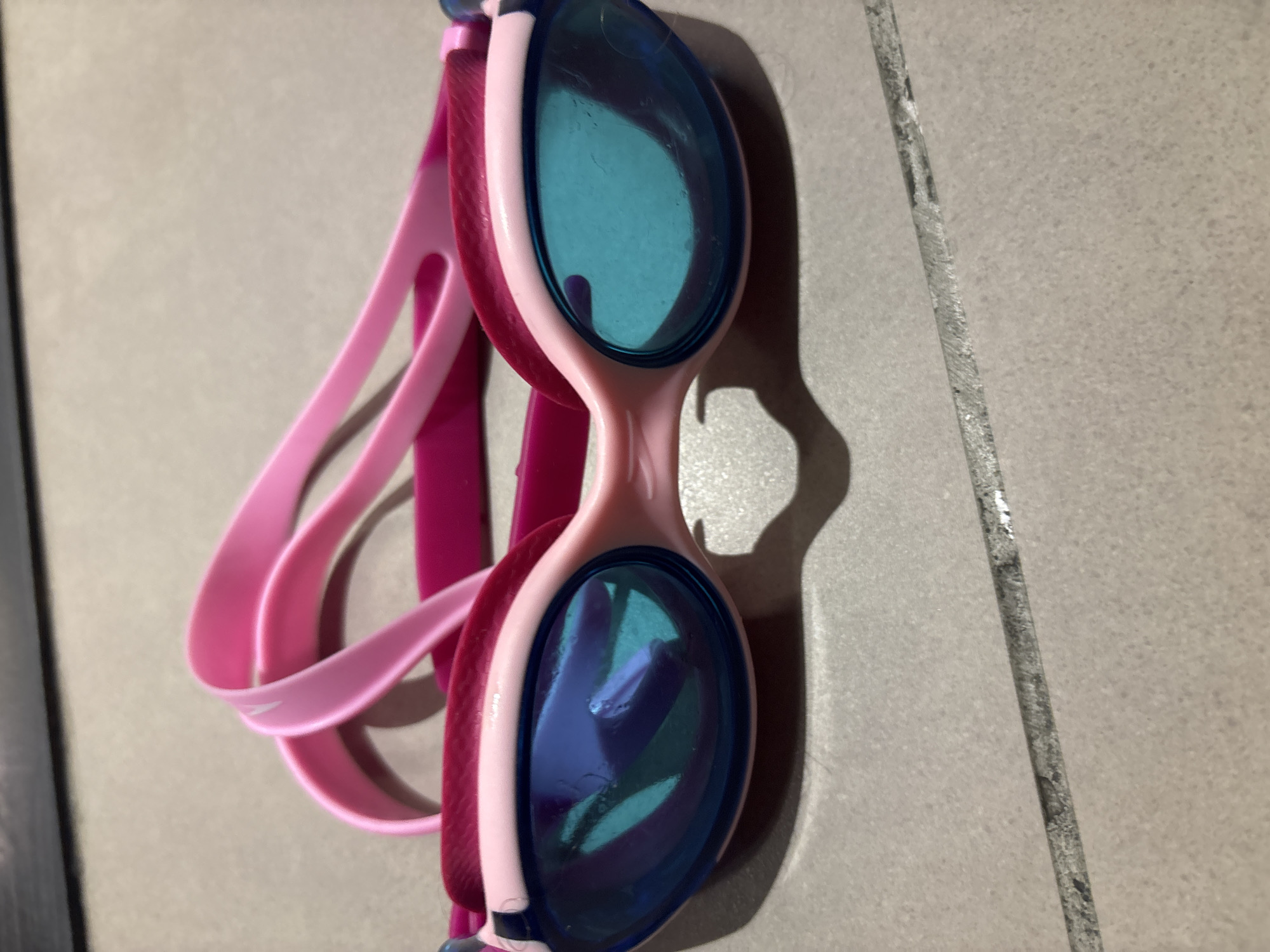 A pair of pink and blue swimming goggles rests on a tiled surface. The goggles have bright blue lenses with a turquoise tint, reflecting the surroundings. The frame of the goggles is primarily pink, with a darker pink band around the lenses. A bright pink strap extends from the goggles, with cutouts for adjustment. The tiled surface appears to be light gray and is laid out in a grid pattern. The background is out of focus, showing a dark horizontal surface above the tiled area. There is a shadow cast by the goggles onto the tiles. No people are visible in the image. The scene is a close-up, eye-level shot of the swimming goggles.
