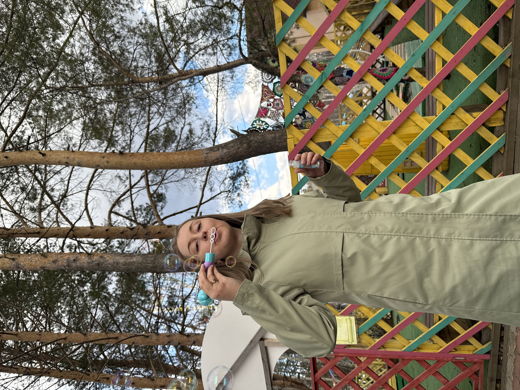 A person in a light green jacket is blowing bubbles in an outdoor location in Unknown, Unknown. The background includes trees and a colorful wooden structure with yellow, green, and pink accents. The sky is visible through the branches of the trees. Bubbles are floating near the person's face.