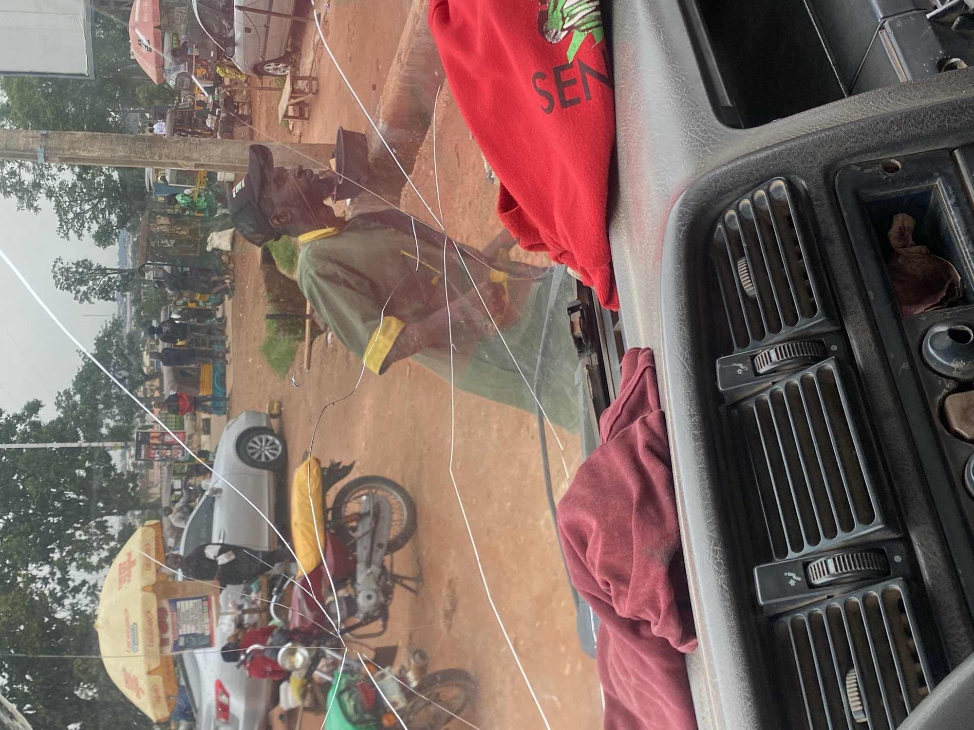 The image depicts a street scene in Gura Top, Nigeria, viewed from inside a vehicle through a cracked windshield. In the foreground, the dashboard of the vehicle is visible, with air vents and some debris. Outside, a man wearing a green shirt and a cap is standing and looking to the right. He is near a red cloth that is draped over the dashboard. Behind him, there are several vehicles, including motorcycles, cars, and what appear to be tricycles, which are common modes of transport in Nigeria. Various stalls and people are also visible in the background, suggesting a busy market or roadside commercial area. The environment is dusty and appears to be an unpaved road. The overall impression is of a typical urban street scene in Nigeria.