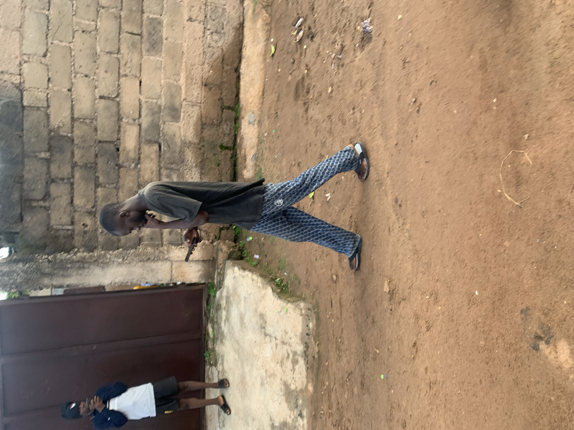 In Gura Top, Nigeria, a young man walks by a brick wall. He is wearing a black t-shirt and blue and white checkered pants. He is holding a phone to his ear with his left hand. To the left of him, another person stands near a large brown gate, also holding a phone. The ground is dirt, and there is some debris scattered around.