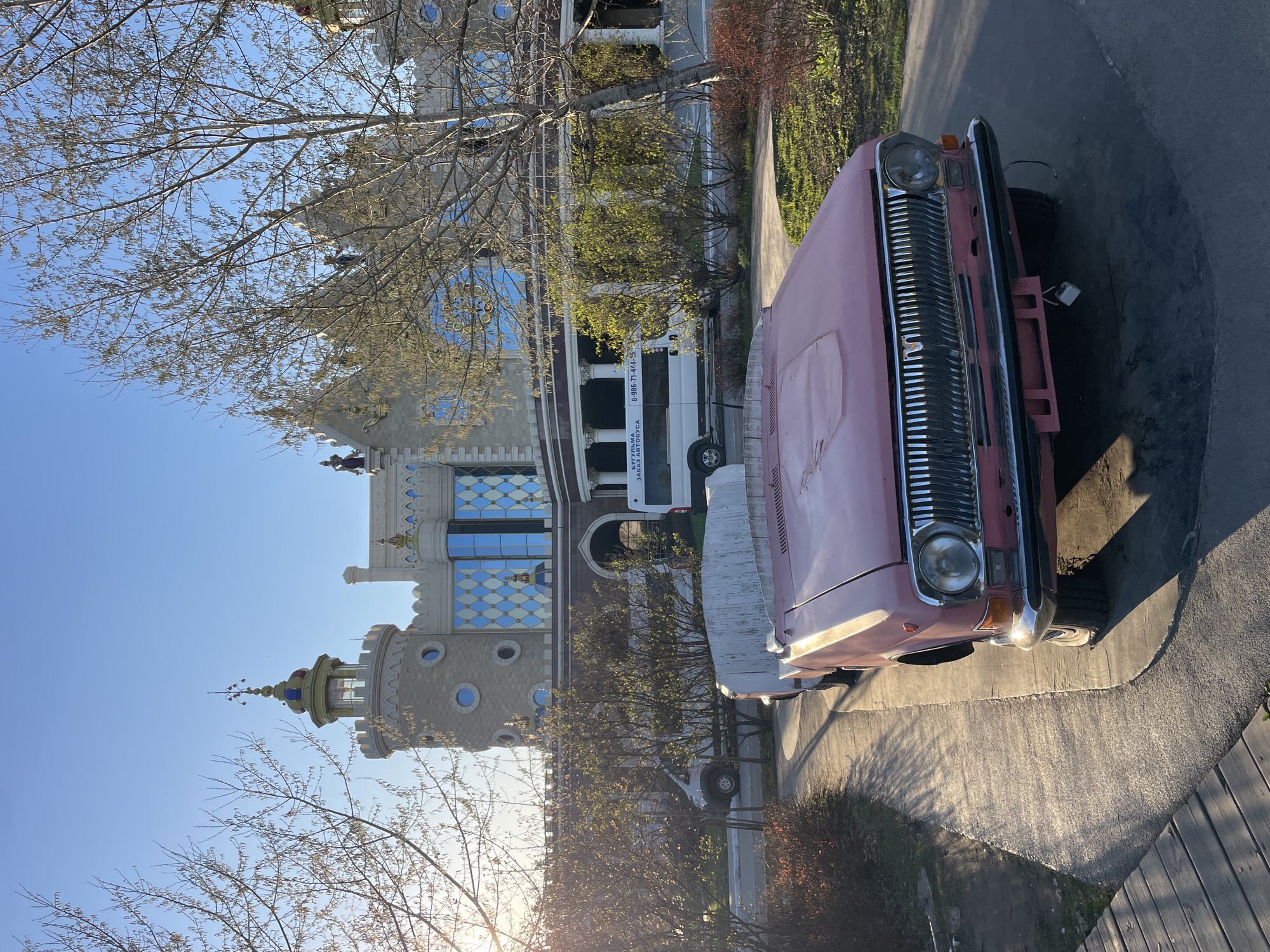 An eye-level, outdoor shot of a pink car parked on the street. In the background is the Kazan Puppet Theater located in the city of Kazan, Russia. A white bus with lettering on the side is parked in front of the theater. The sky is blue and a tree in the foreground is partially covering the theater.
