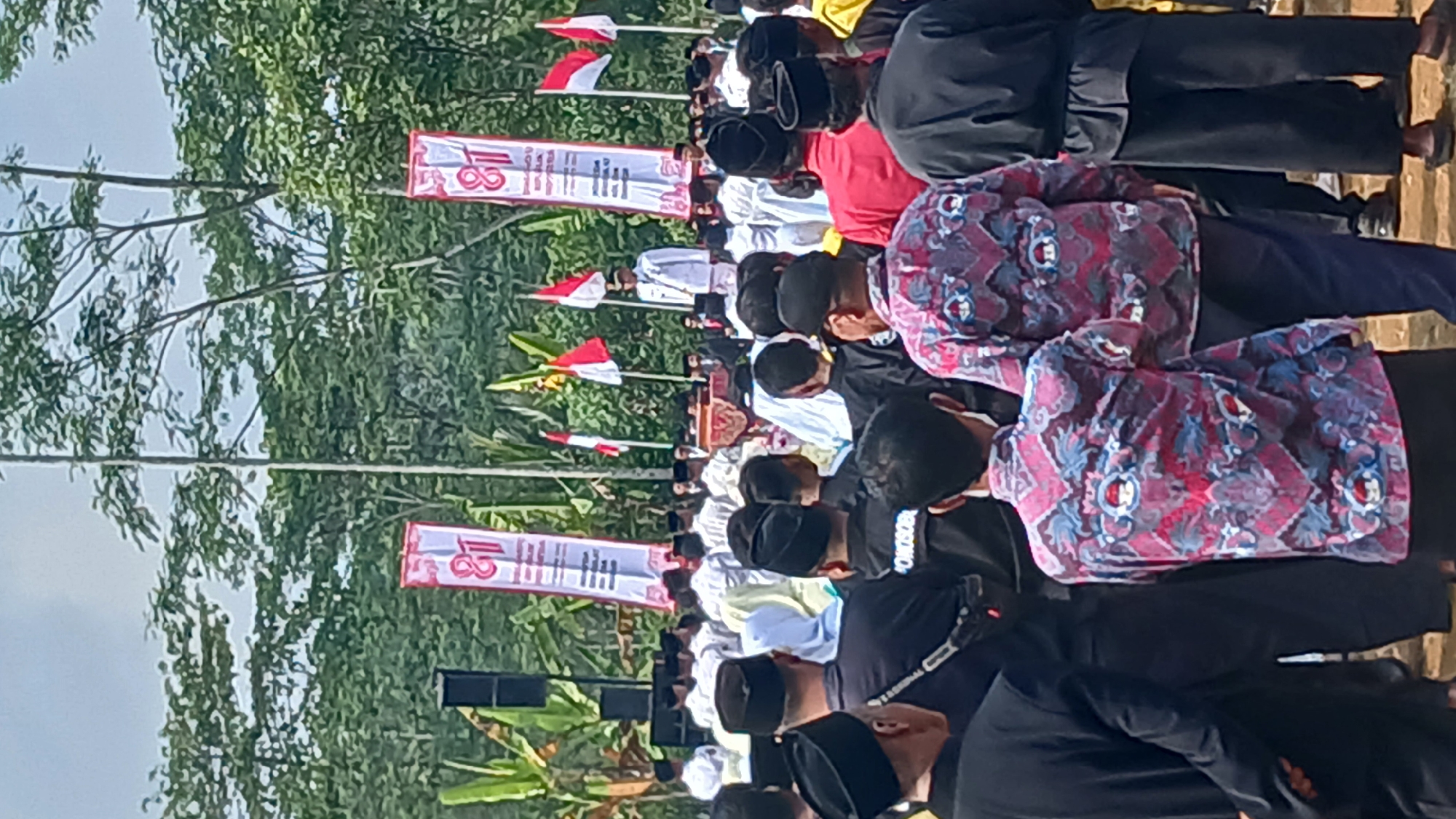 The image captures an outdoor gathering in Kepil, Indonesia, likely a public event or ceremony. A crowd of people, predominantly male, are seen from behind, many wearing traditional Indonesian attire such as peci hats and batik shirts. In the background, several tall red and white banners with the number "81" are prominently displayed, suggesting a celebration of an 81st anniversary or event. Red and white Indonesian flags are also visible. A man in a white suit stands on a raised platform. The environment appears to be rural, with lush green trees and foliage forming the backdrop. The ground is unpaved, indicating an outdoor, possibly natural setting.
