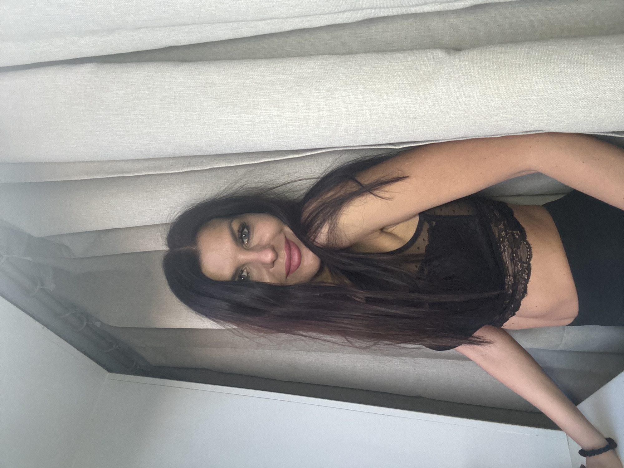The image captures a headshot and upper body of a woman indoors, potentially in Chaikovsky, Russia. The woman, with long, dark hair, green eyes, and pink lipstick, is smiling faintly as she looks towards the camera. She is wearing a black top with intricate lace detailing, possibly a bralette, and black bottoms. A black scrunchie or bracelet adorns her left wrist.
The background consists of a light-colored, possibly off-white or light gray, textured fabric with horizontal folds, resembling curtains, blinds, or an upholstered surface like a headboard or sofa back. A section of a plain white wall is visible in the lower left corner. The lighting appears soft and diffused, suggesting natural light, though the exact time of day or outdoor weather cannot be determined. The woman is positioned lying down, with the image rotated so she appears to be looking upwards. No text is visible in the media file.